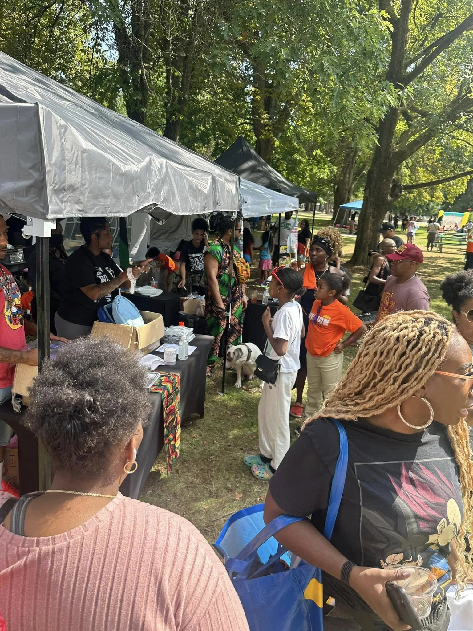 Back To The Bell event in a park, with vendor stalls under tents, trees providing shade, and a bounce house in the background.