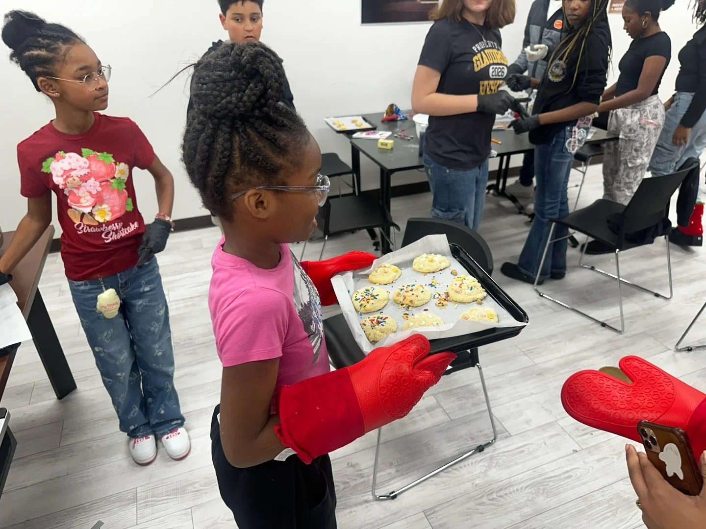 A girl wearing safety goggles and red oven mitts holds a tray of decorated cookies baking at The Junior Bakers Club