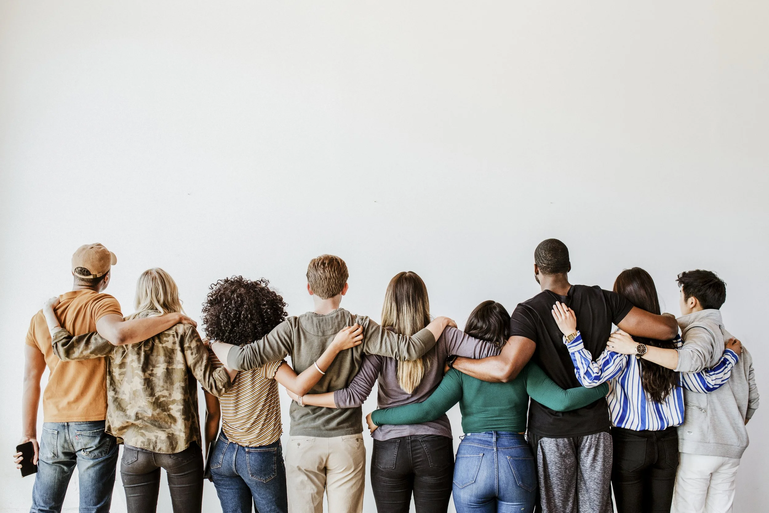 Group of diverse people standing with their arms around each other, facing away from the camera against a plain white wall.