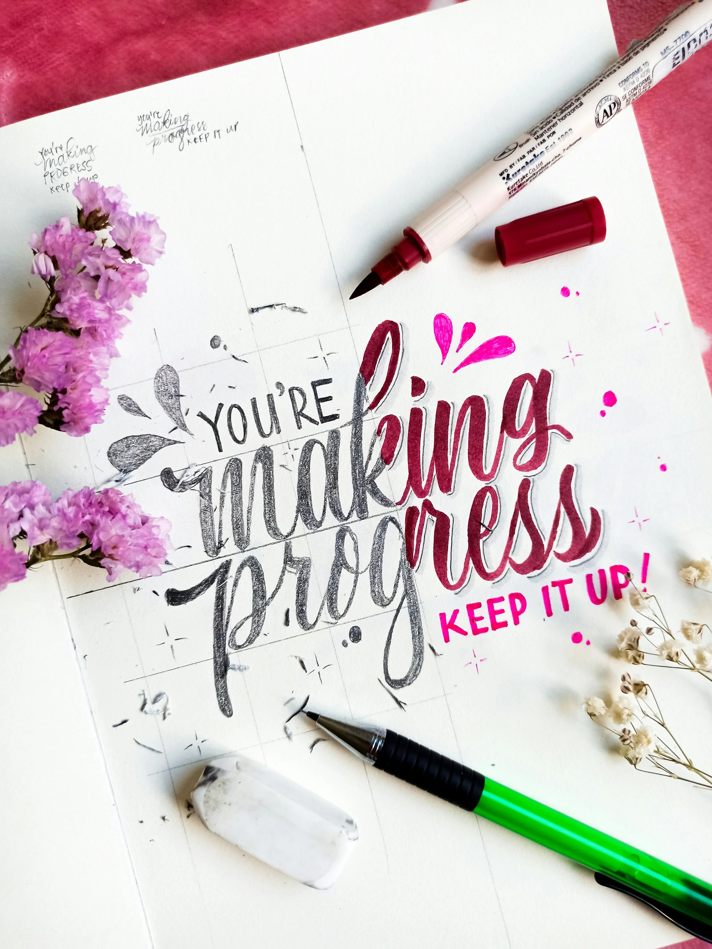 Hand-lettered text reads, "YOU'RE MAKING PROGRESS KEEP IT UP!" in a sketchbook, adorned with flowers, a red marker, an eraser, and a mechanical pencil on a pink background.