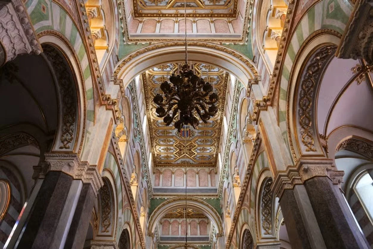 Interno di una chiesa o di un palazzo storico, con soffitto decorato, grandi archi e lampadario antico in metallo