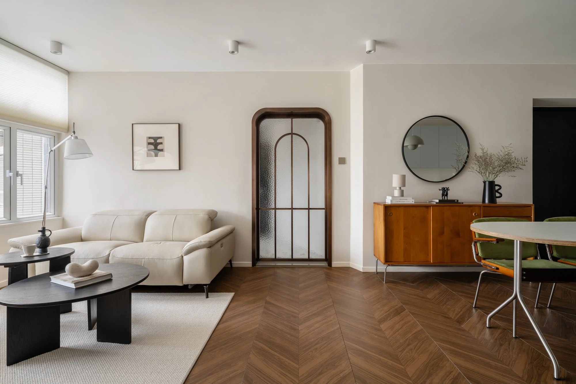 Modern living room with white sofa, black coffee table, wooden sideboard, round mirror, and dining table with green chairs, wood flooring, and minimal decor.
