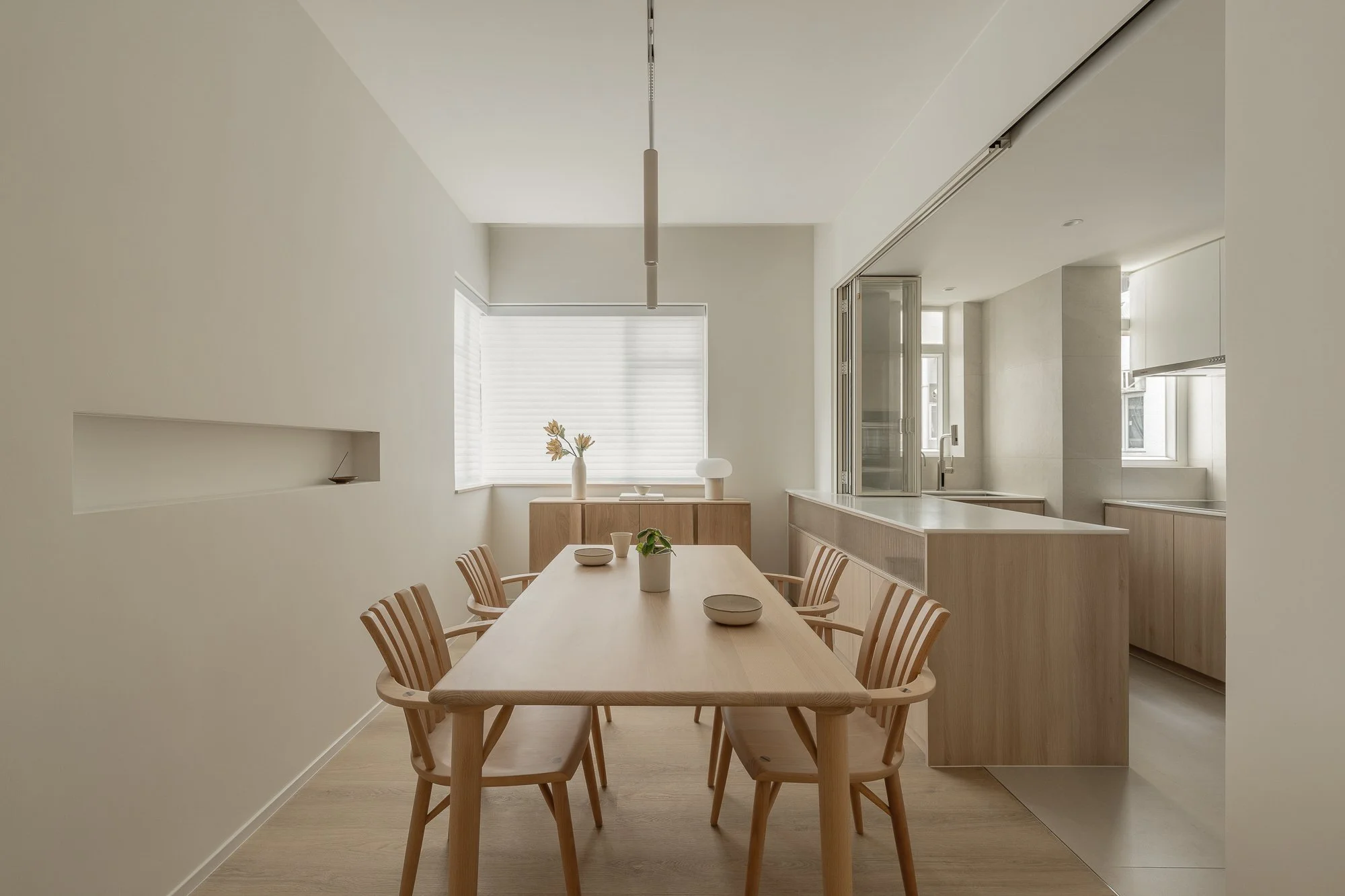 A dining room with a wooden table and chairs, a sideboard with a vase of flowers, a bowl, and a lamp, with large window blinds and a kitchen window in the background.