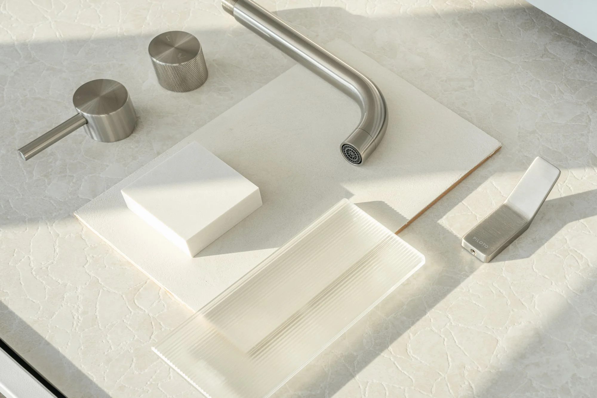 A modern kitchen sink faucet with a matte stainless steel finish, alongside two matching control knobs and a silver utility lighter, all arranged on a light-colored marble countertop with a beige textured background.