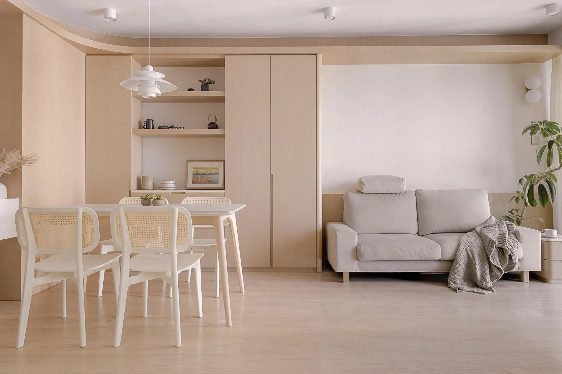 Minimalist living room with a dining area and a beige sofa, light wood flooring, built-in shelving with decor, and a potted plant.