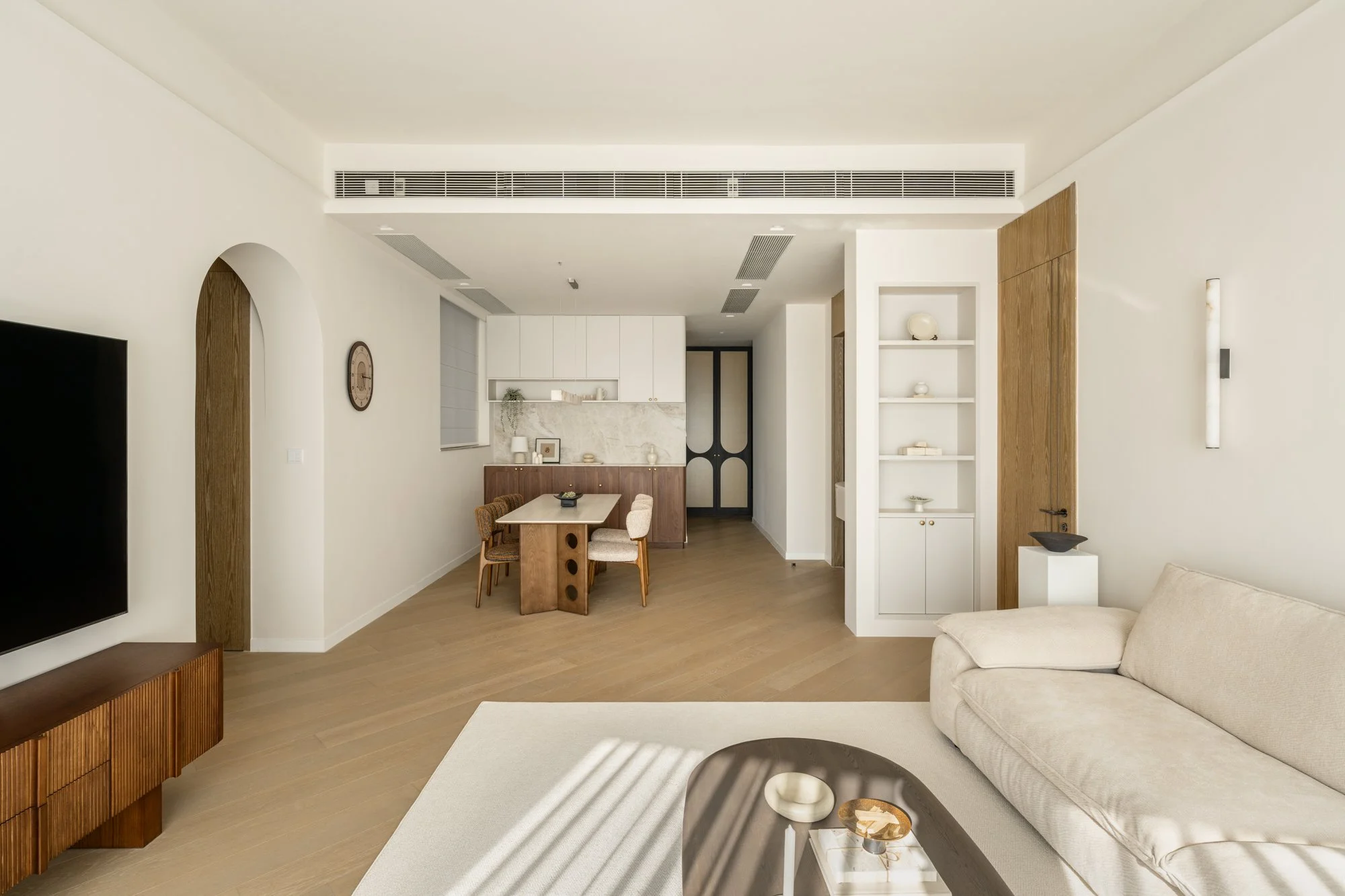 Modern living room with light-colored walls, a beige sofa, a white and wood TV stand, a round coffee table with candles, and a dining area with a wooden table and six chairs. The space features wooden flooring and built-in shelves, with a minimalist aesthetic.