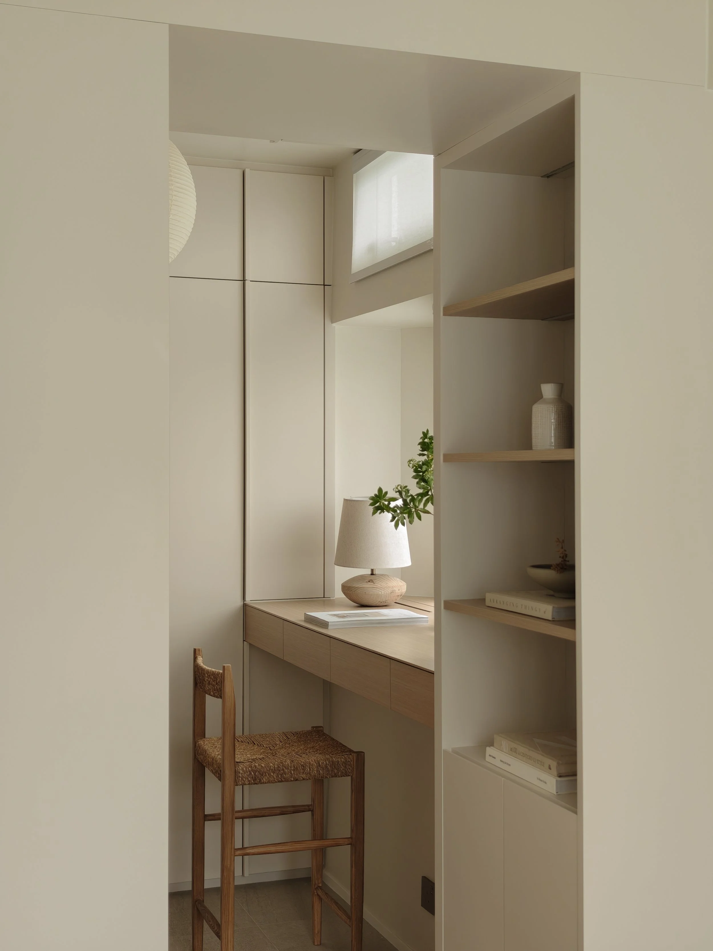 A minimalist interior space with a built-in white bookshelf, a small wooden desk, and a wooden chair with a woven seat. There is a white lamp, a small plant, and a few books and decorative items on the shelves. The space is well-lit with natural light coming from a window above the desk.