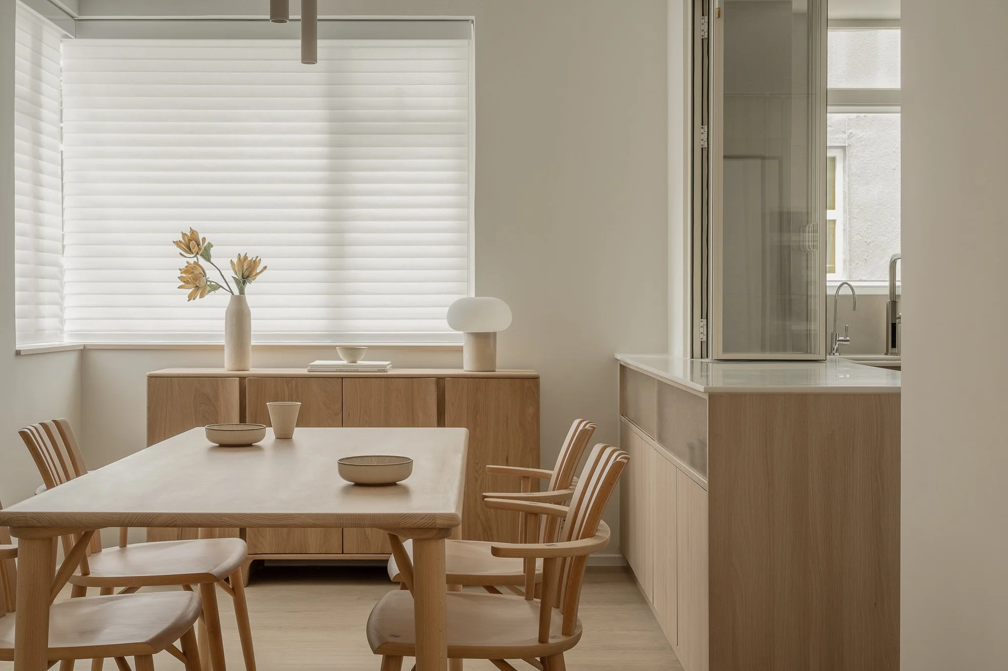 A dining room with a wooden table and chairs, a sideboard with a vase of flowers, a bowl, and a lamp, with large window blinds and a kitchen window in the background.