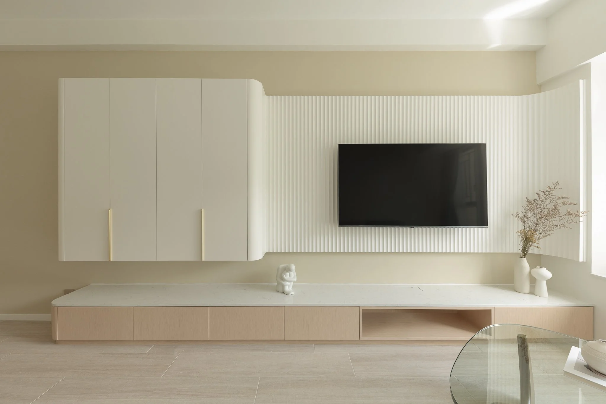 Modern living room with a wall-mounted flat-screen TV, white textured wall panels, beige and white storage units, and decorative vases with dried plants.