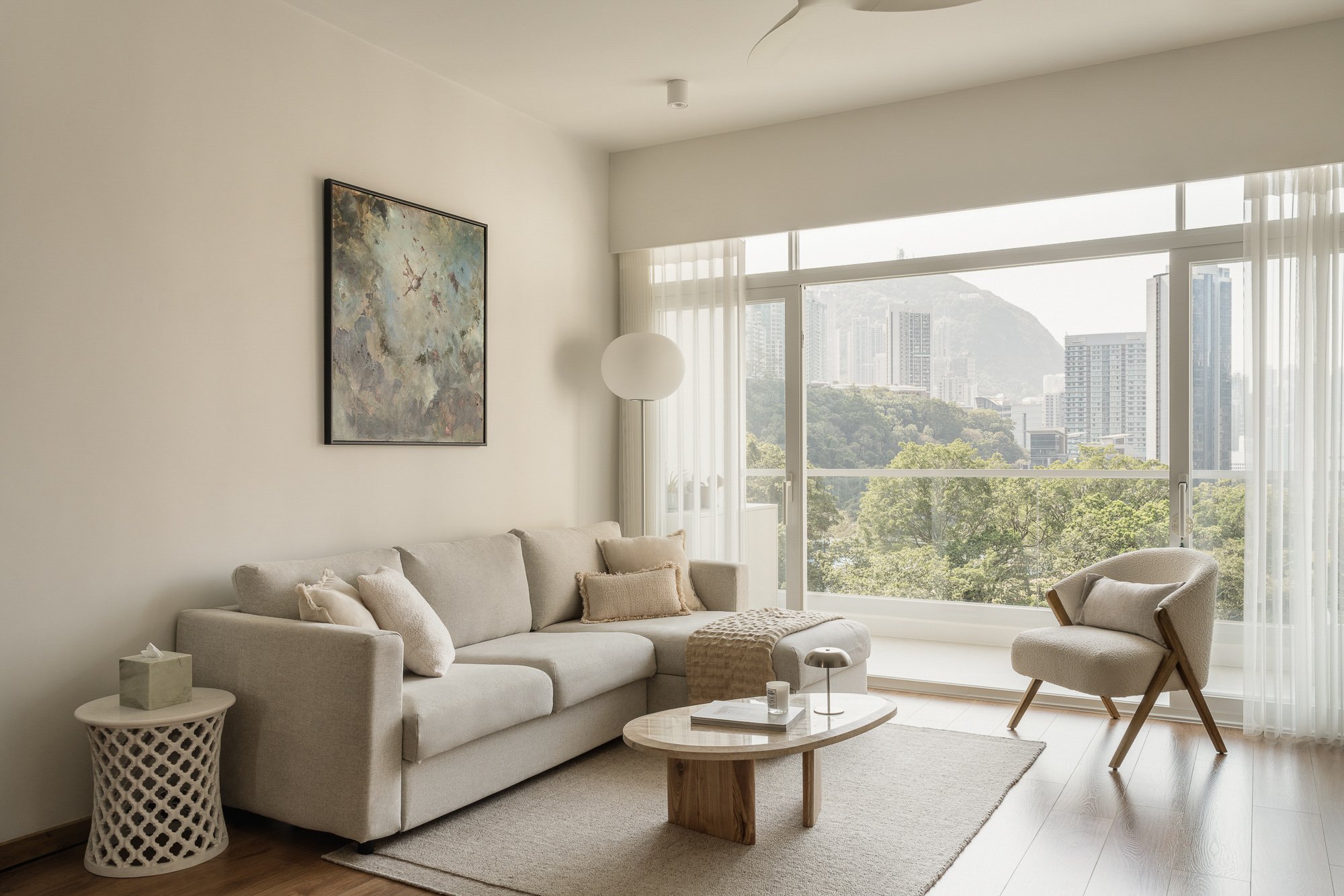 A modern living room with a large beige sofa, a small round marble-top coffee table, and a white armchair. There is a white lattice side table and a floor lamp with a globe-shaped shade. The room has floor-to-ceiling windows with sheer curtains, allowing natural light and a view of city buildings and a hillside.