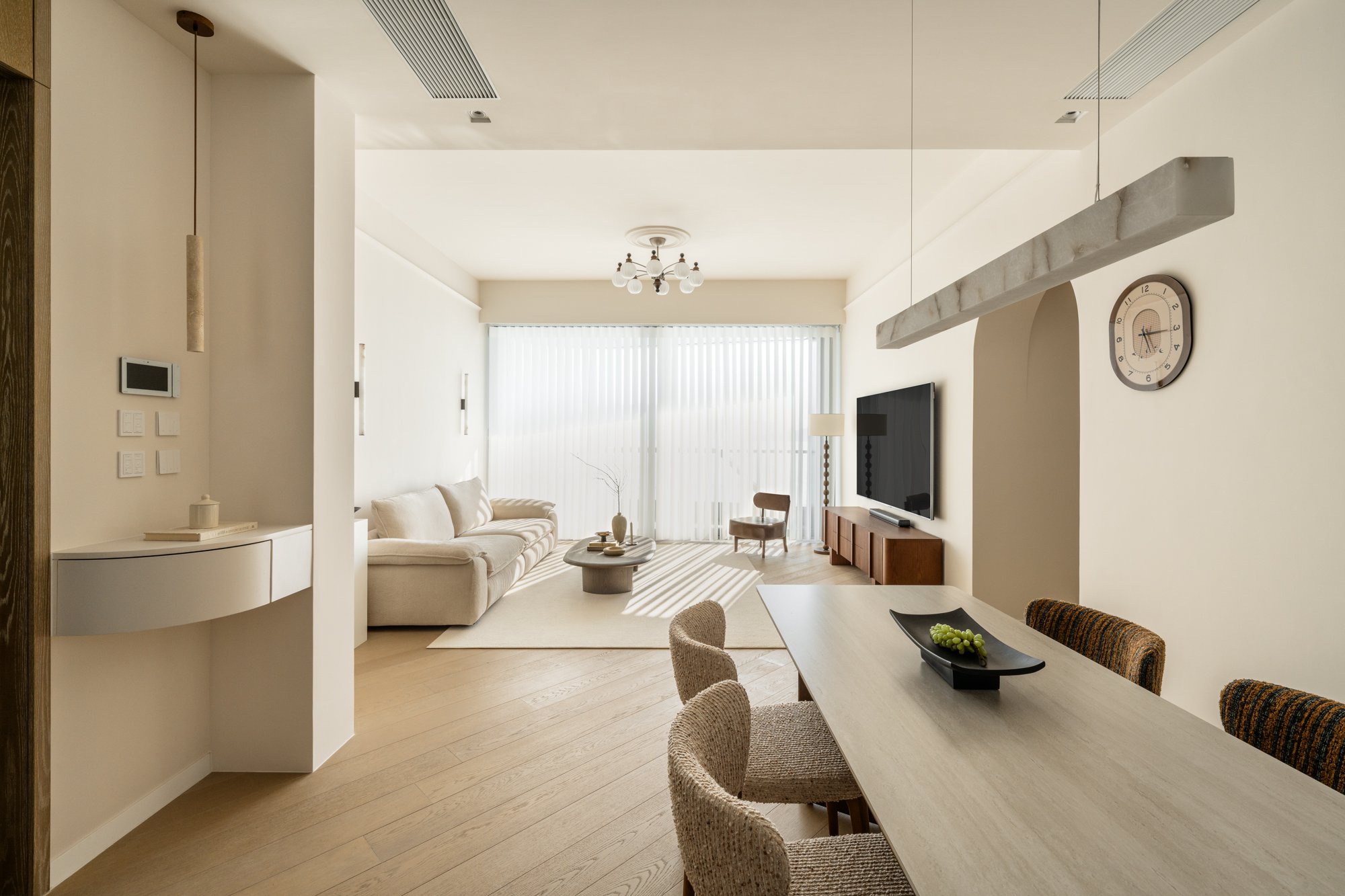 A modern living room with a white sofa, a wooden TV stand, a flat-screen TV, and a round coffee table. Large windows with vertical blinds, a chandelier, and a wall clock are also visible.