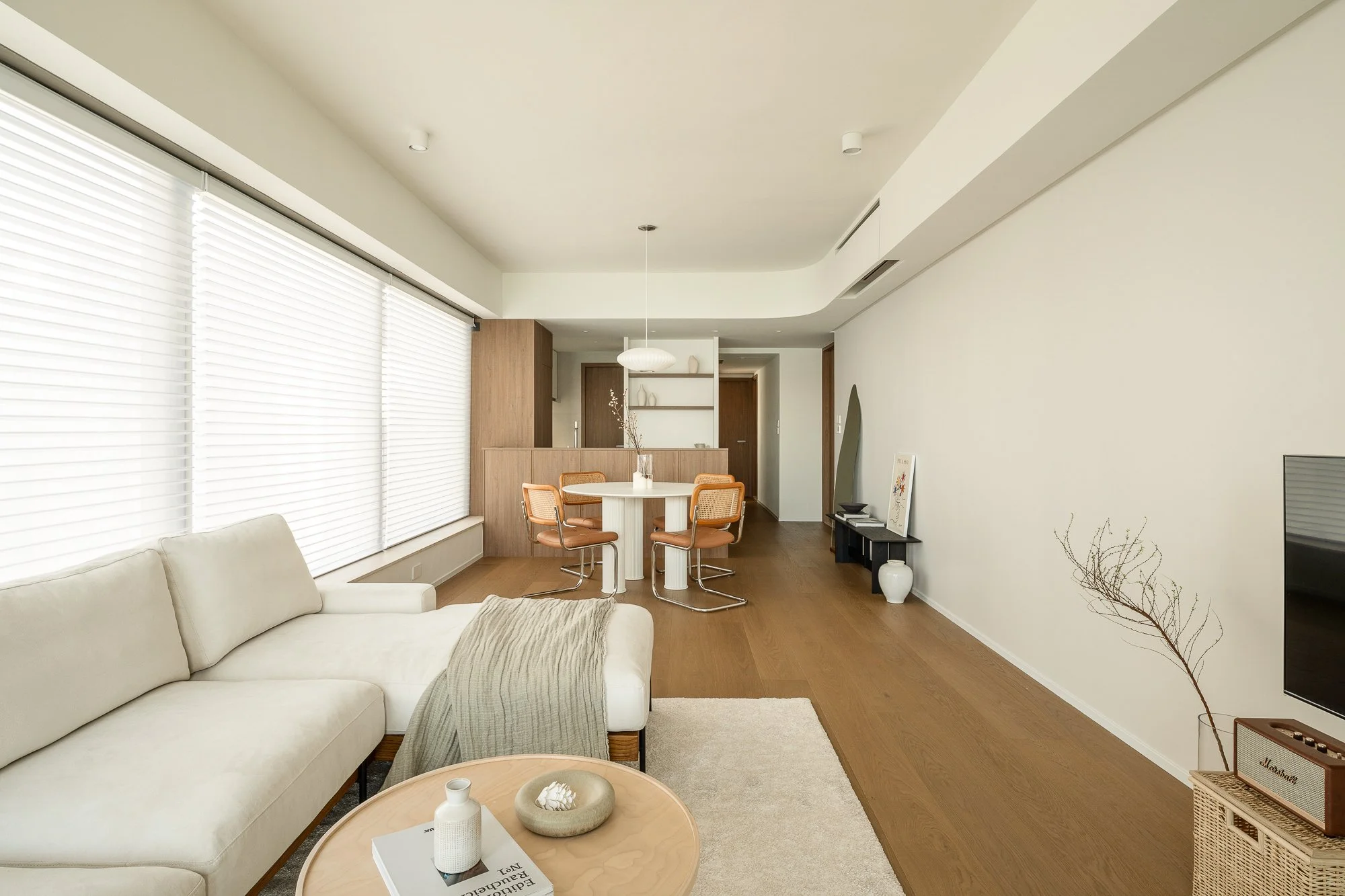 Living room with white sofa, wooden floor, circular dining table with four chairs, large window with blinds, TV, minimalist decor including a mirror, artworks, and vases.