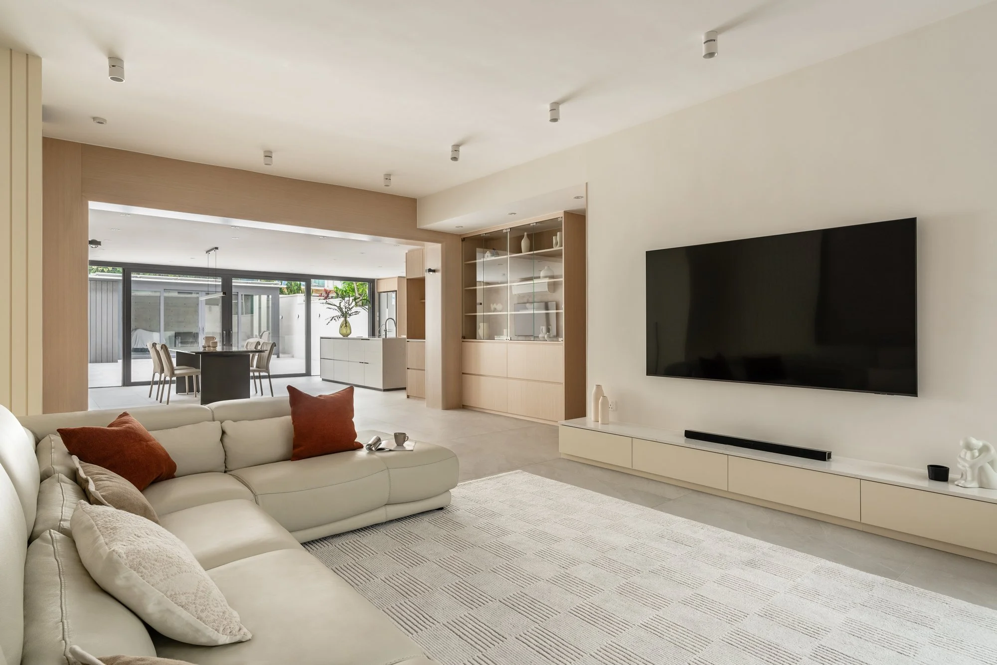 Modern living room with a beige sectional sofa, decorative pillows, wall-mounted flat-screen TV, white media console, and a view of the dining area and kitchen through an open layout.