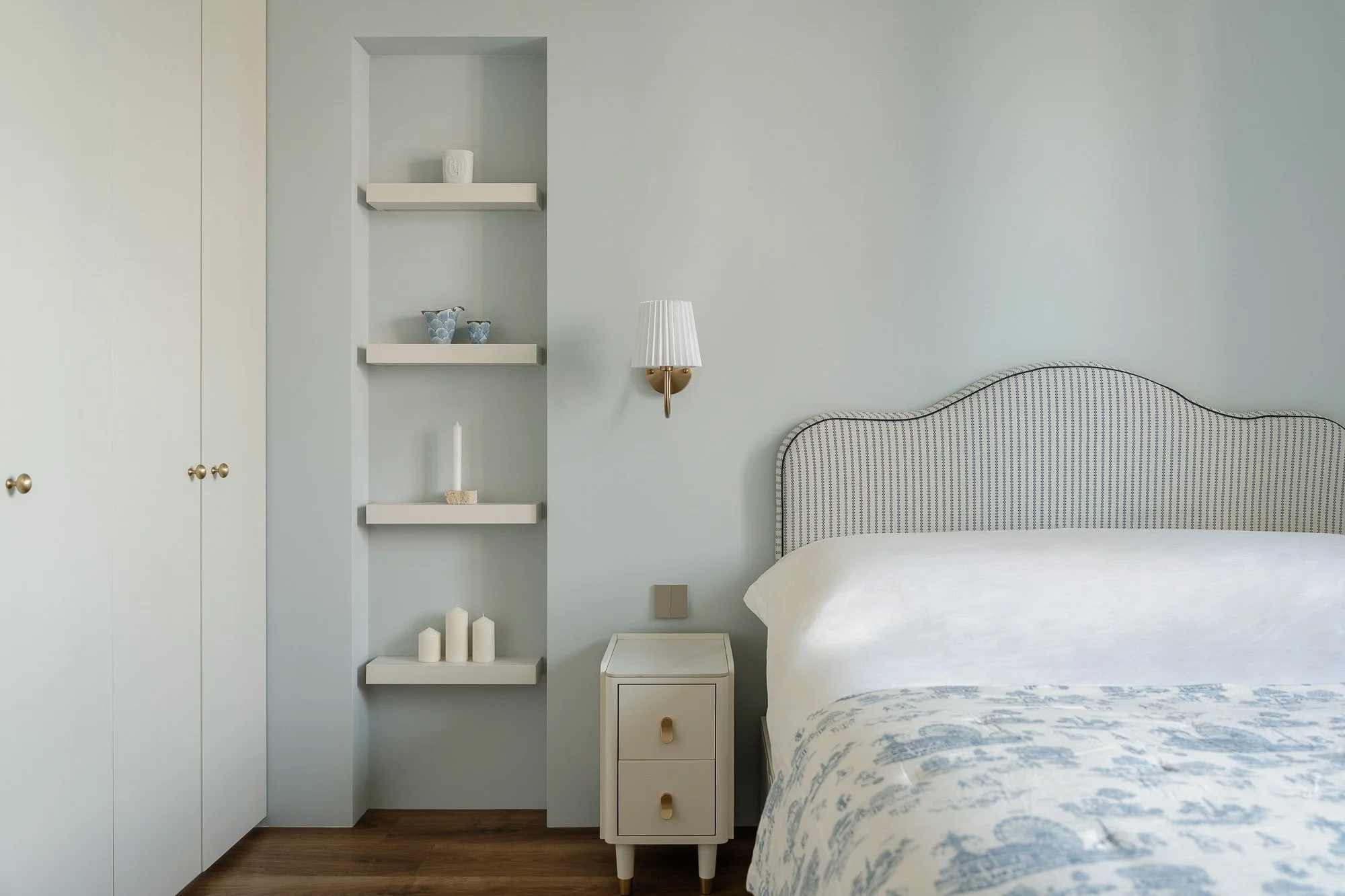A bedroom with a light blue wall, a white bed with blue patterned bedding, a white nightstand, wall-mounted light, and decorative shelves with candles and small blue vases.