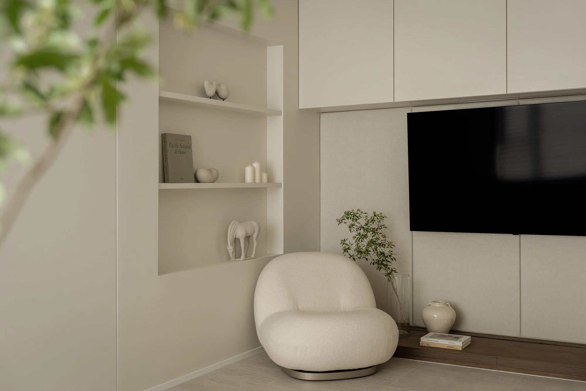 A modern, minimalist living room corner featuring a white, curved armchair, a small potted plant, a white vase, and a white bookshelf with decorative objects and books, against a beige wall.