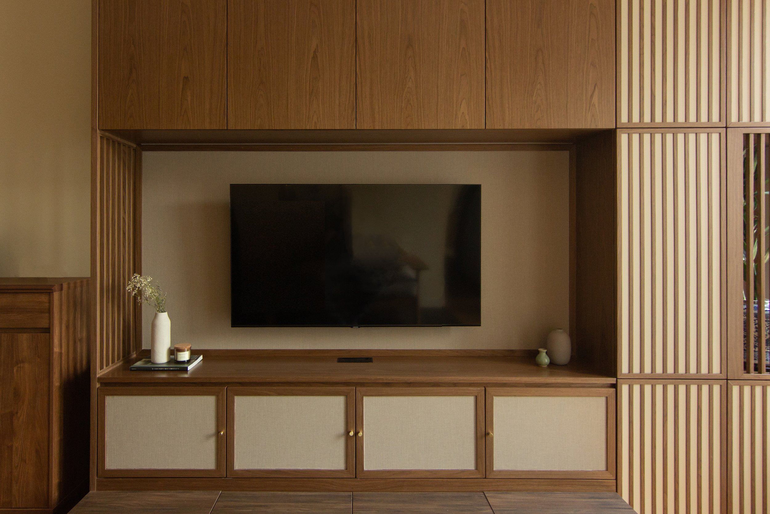 A wooden wardrobe with sliding doors, one revealing hanging white shirts and a small drawer at the bottom, and a top shelf with a small potted plant and a book or tablet.