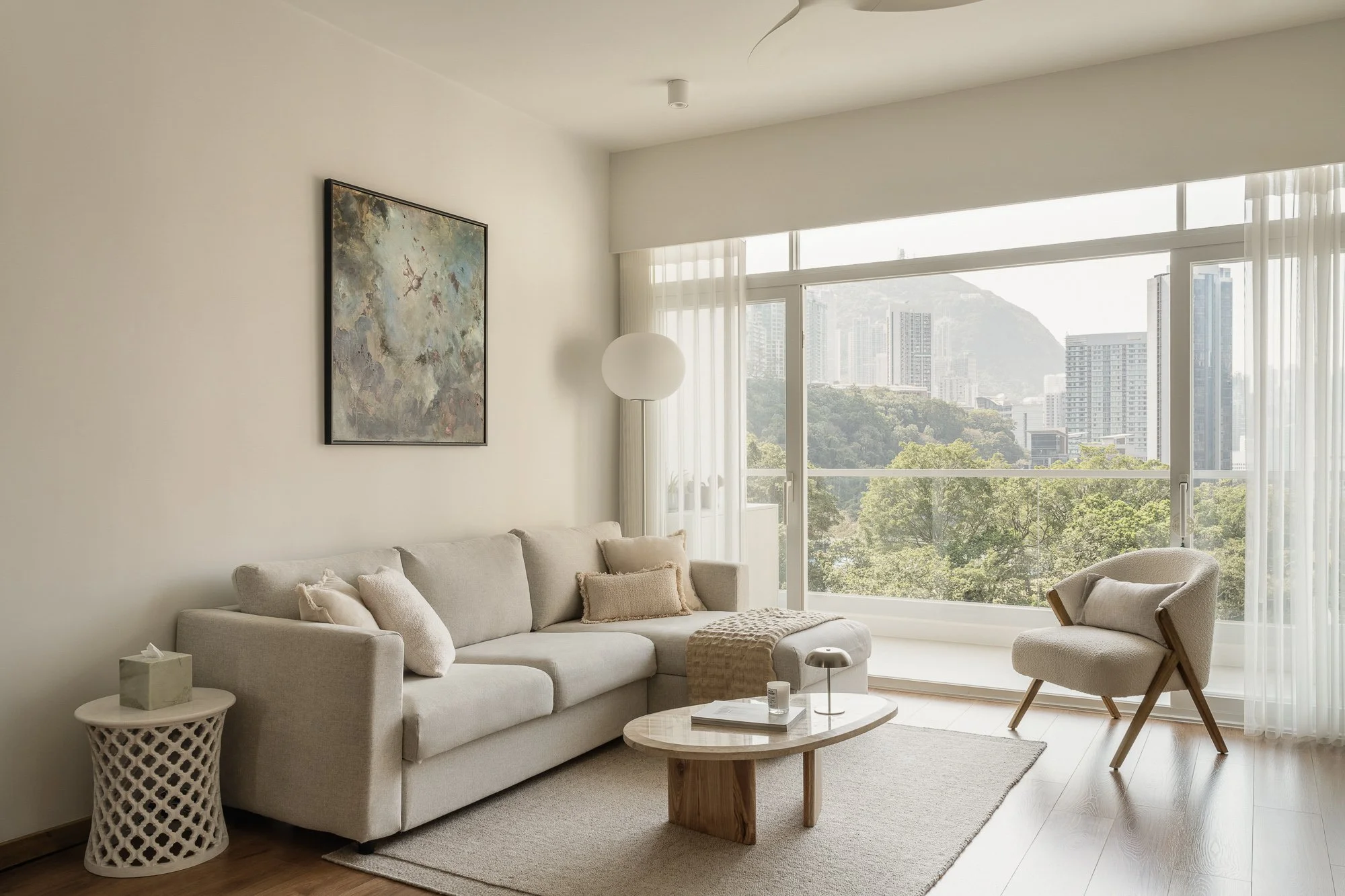 Modern living room with large windows, white walls, beige sofa, wooden coffee table, and a white armchair. There are sheer curtains and a cityscape view with tall buildings and green trees outside.
