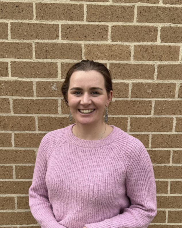 A woman with brown hair tied back, smiling, wearing a pink knitted sweater and earrings, standing in front of a brick wall.