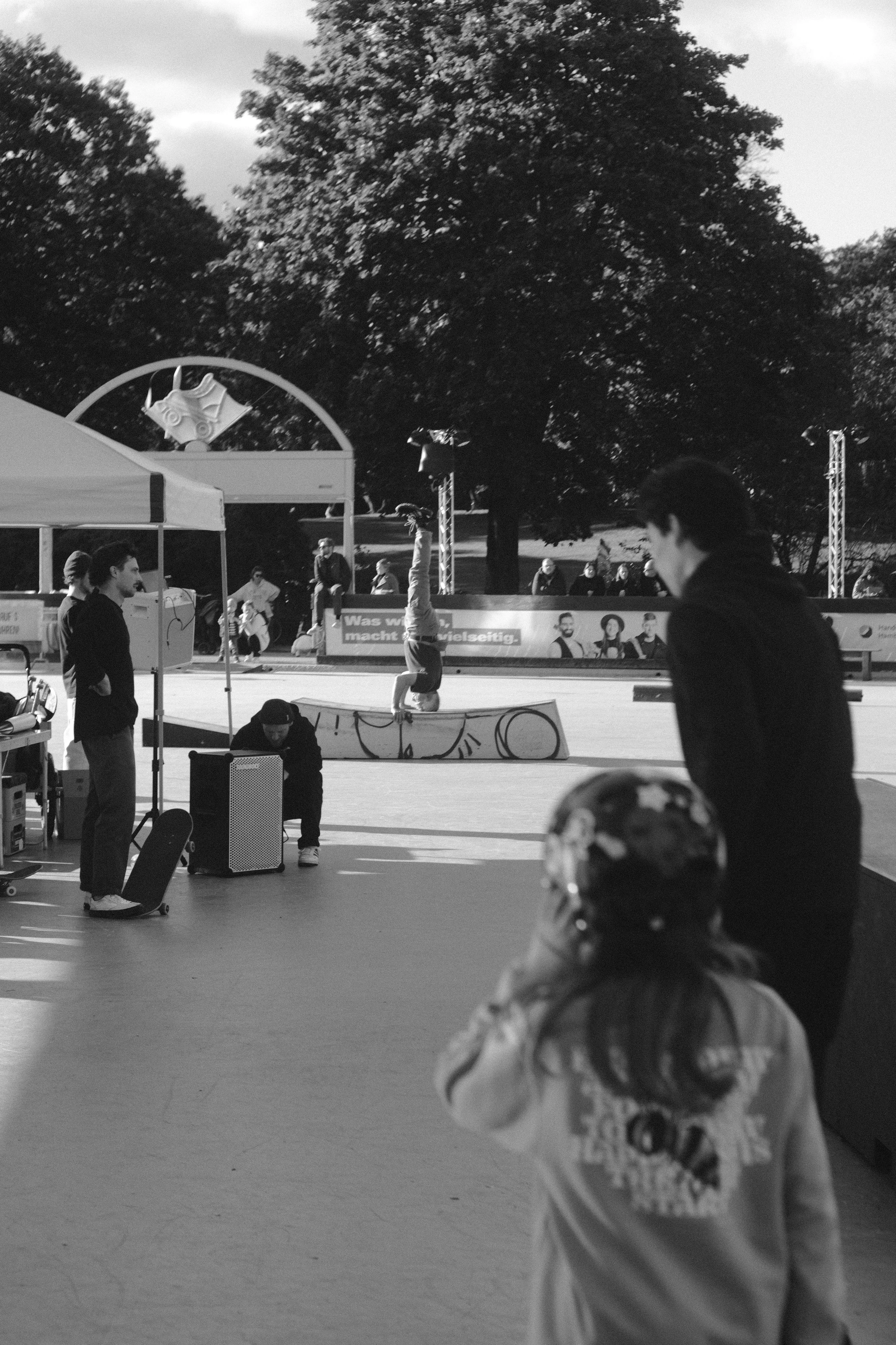 Kinder beim Skaten und BMX-Fahren auf einer Skateanlage im Freien, umgeben von Bäumen, an einem Tag mit bewölktem Himmel.