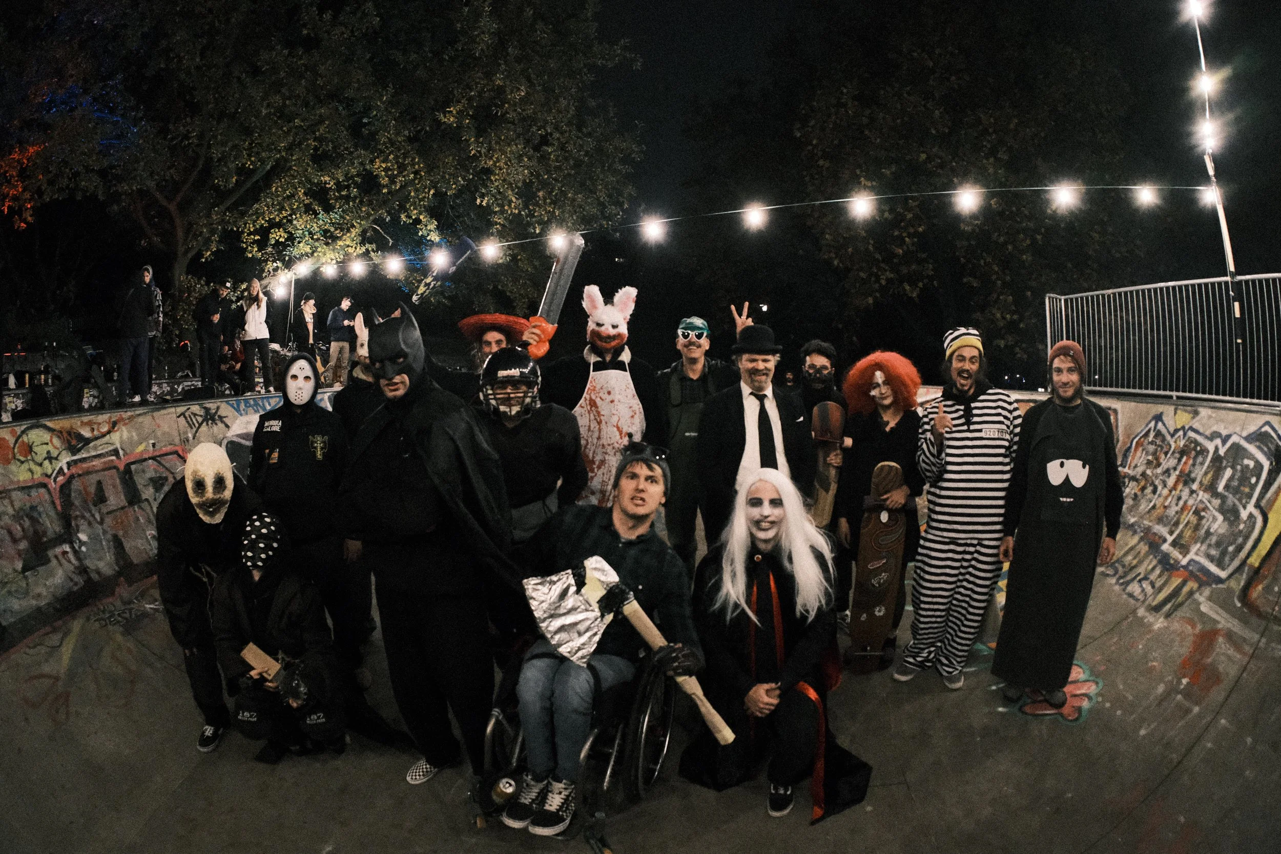 Menschen in Halloween-Kostümen bei einer nächtlichen Veranstaltung in einem Graffiti-unterführten Skatepark, einige mit Masken und Verkleidungen, in einer lockeren Gruppe, unter Lichterketten