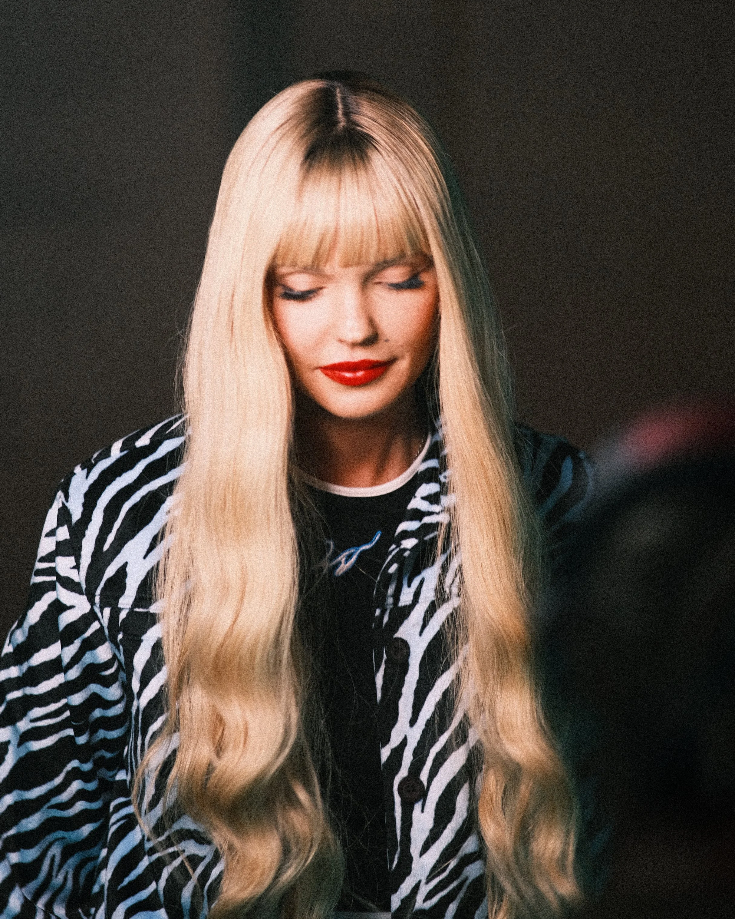 Junge Frau mit langen blonden Haaren und roten Lippen in einem schwarzen und weißen Zebra-Muster-Shirt, nach unten schauend, vor dunklem Hintergrund.