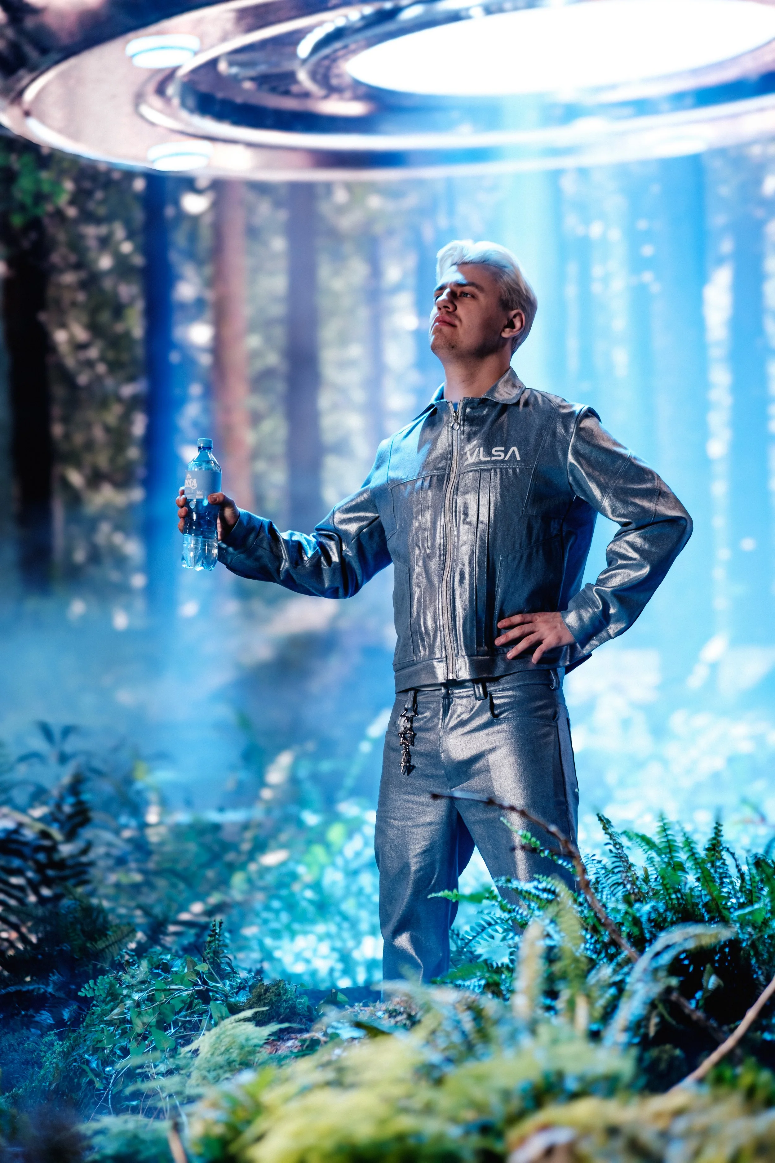 Ein Mann mit silberner Jacke und Hose steht in einem surrealen Wald mit bläulichem Licht, hält eine Wasserflasche in der Hand und schaut nach oben.