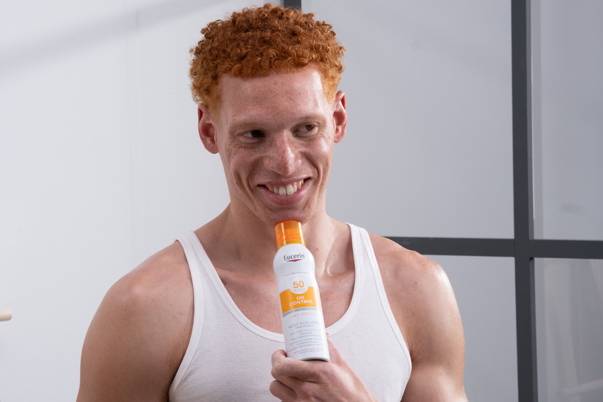 Ein junger Mann mit roten lockigen Haaren trägt ein weißes Tanktop und hält eine Sonnencreme mit SPF 50, während er lächelnd in die Kamera blickt.