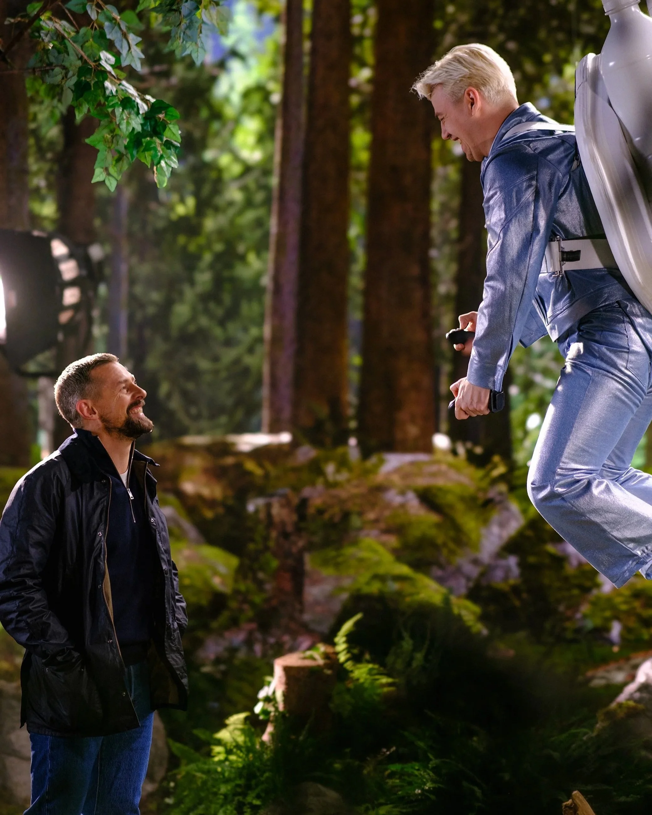 Zwei Männer im Wald, einer kniet und lächelt, der andere steht mit einem Rucksack, beide lachen und interagieren fröhlich, umgeben von grünen Bäumen.