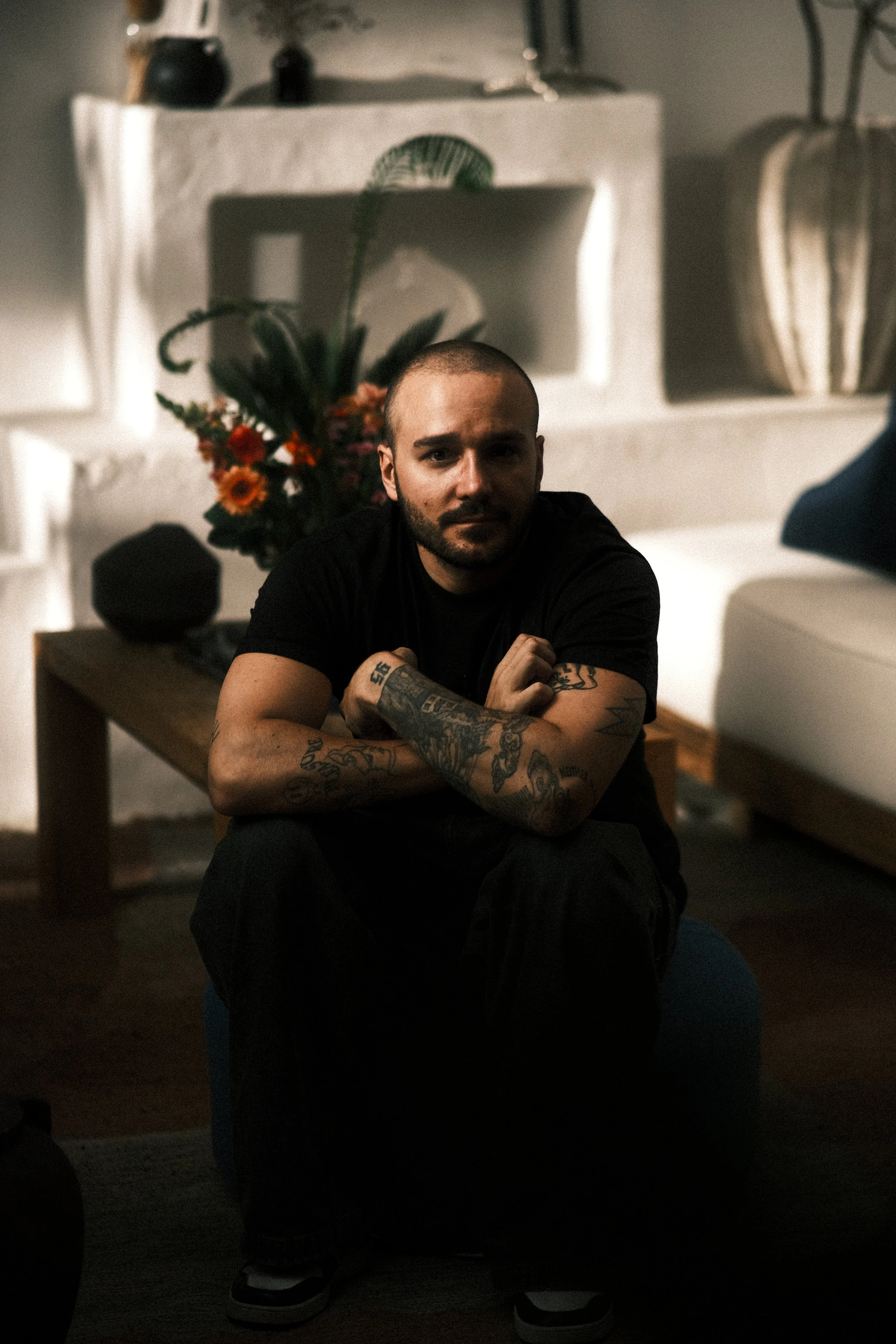Mann mit Tattoos sitzt auf einem Stuhl in einem modernen Wohnzimmer, im Hintergrund eine weiße Kaminumgebung mit Blumenarrangement.