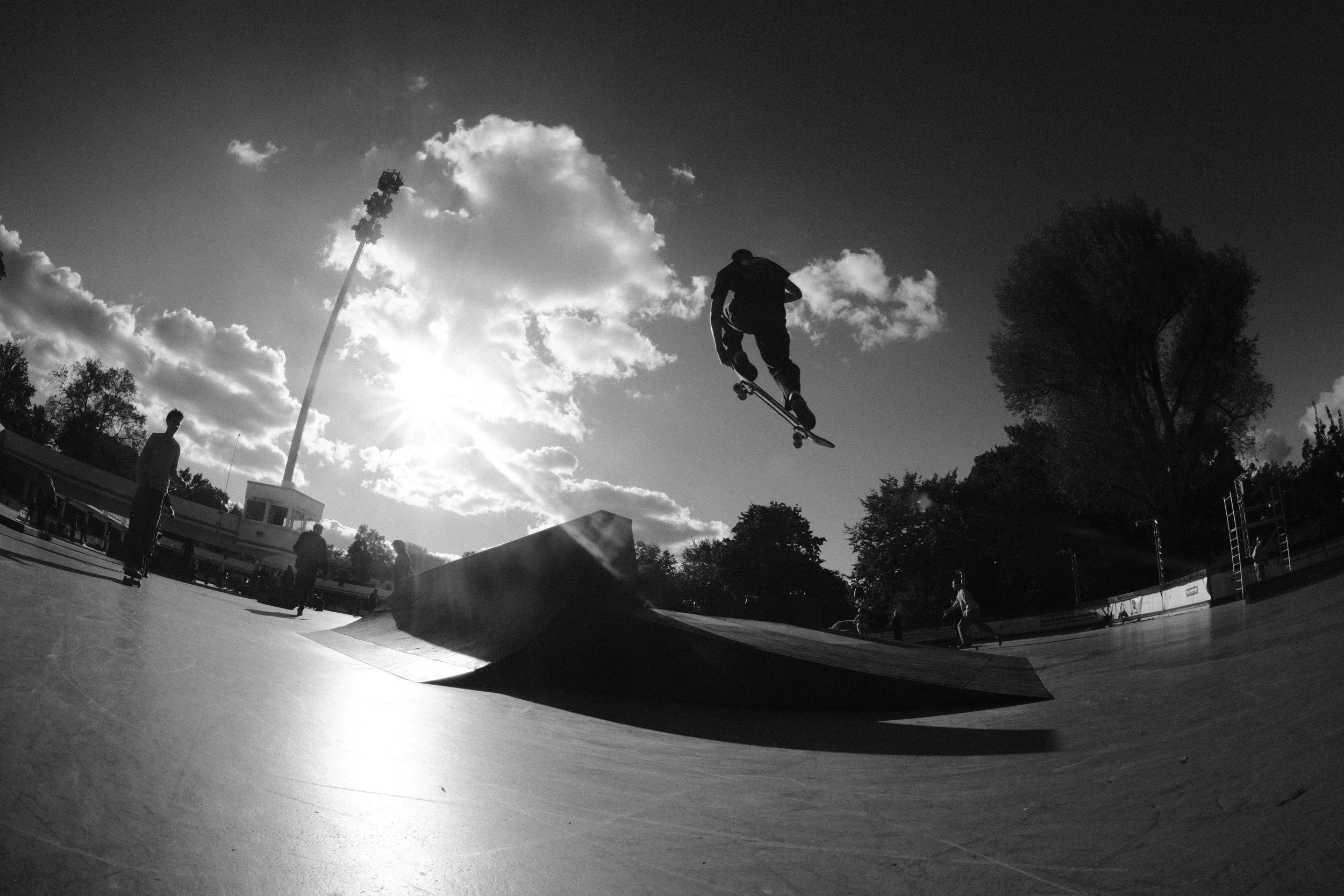 Skateboarder springt in einem Skatepark bei Sonnenlicht, im Hintergrund sind Wolken, Bäume und weitere Personen.