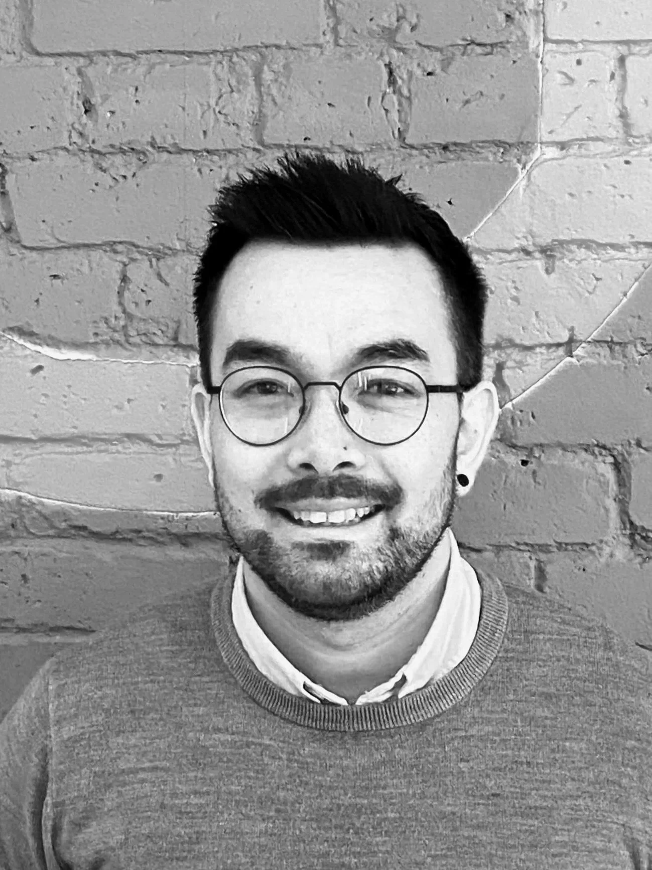 A man with short hair and a beard, wearing a knitted sweater, looks at the camera in front of a circular mirror.