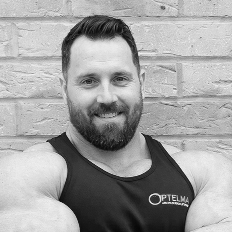 A man with a beard and short hair, smiling at the camera, wearing a sleeveless athletic shirt with a logo that says 'Optelma.' He is standing against a brick wall.