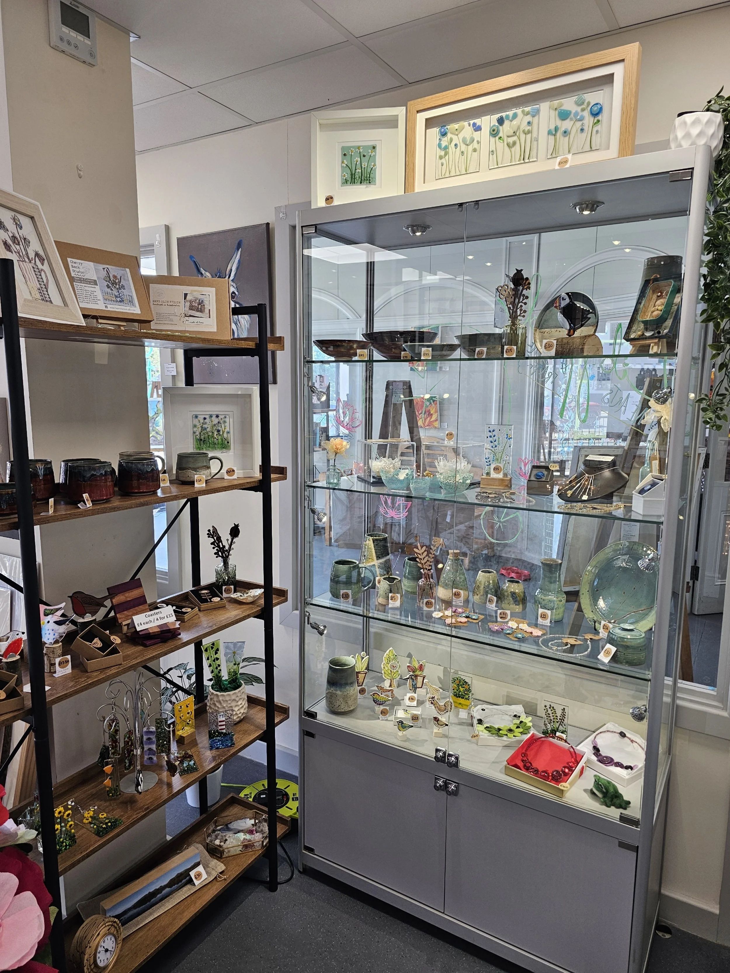 A display case filled with colourful glass art and jewellery, surrounded by additional small art pieces and crafts on shelves, in the gallery.