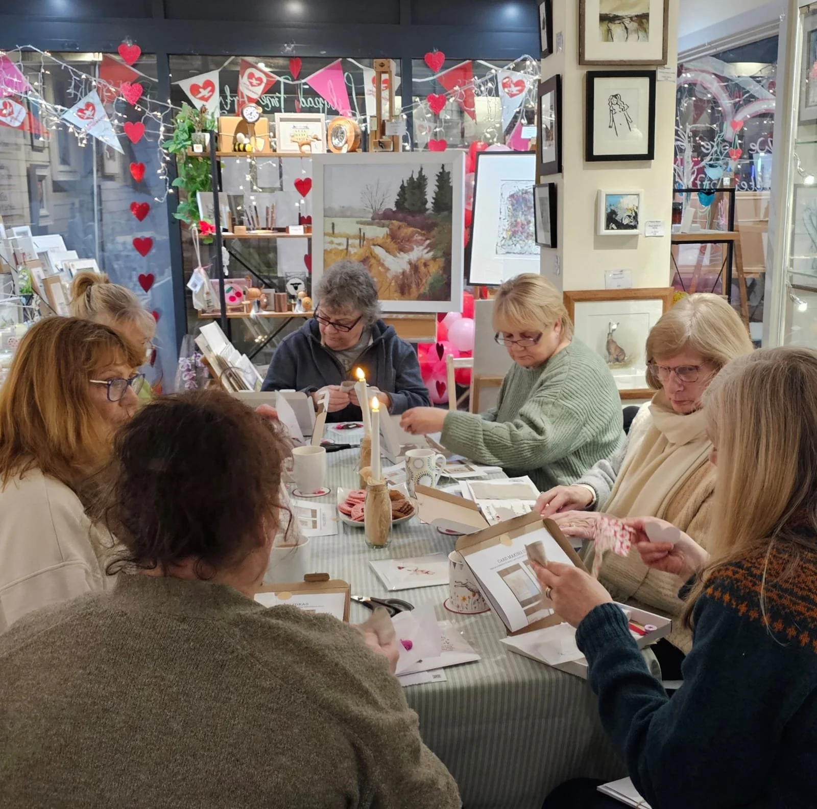 A group of women sitting around a table in a cozy, decorated room, engaged in a craft or workshop activity. The room features framed artwork on the walls, festive Valentine's Day decorations, and a variety of crafting supplies on the table.