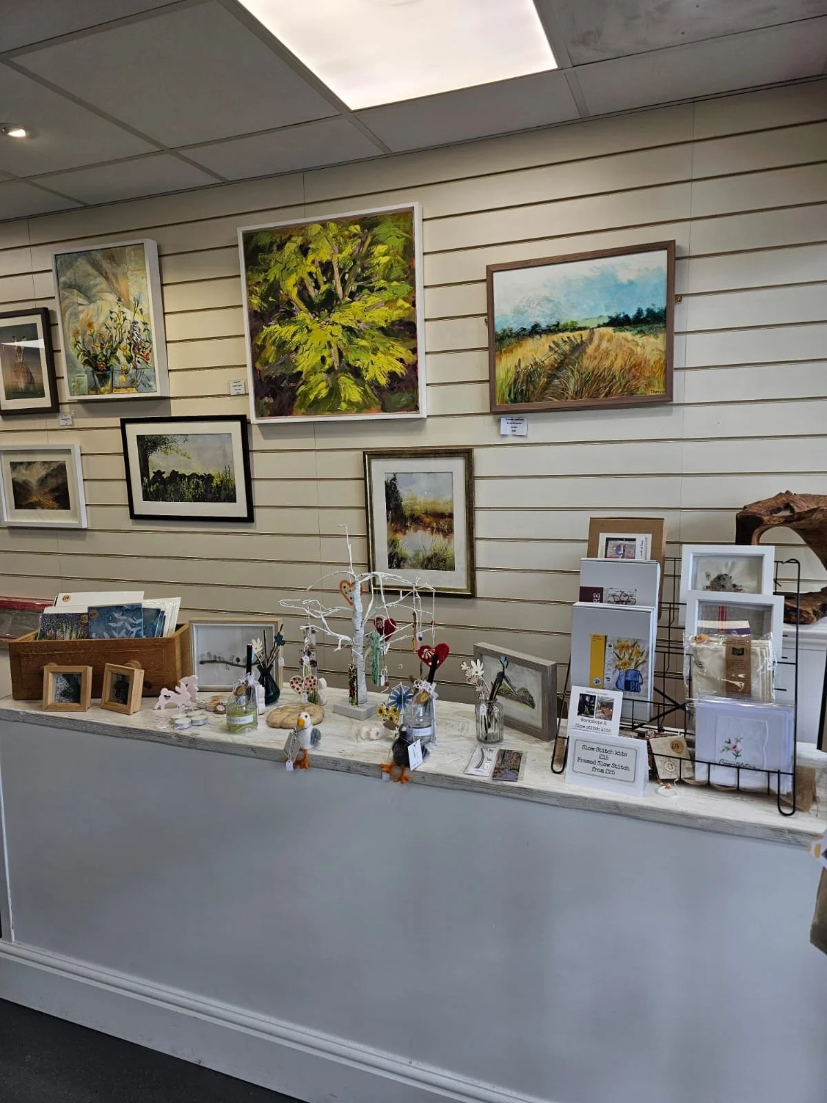 Wall display of various landscape paintings and framed art pieces, with a table in front showcasing handmade crafts and greeting cards.