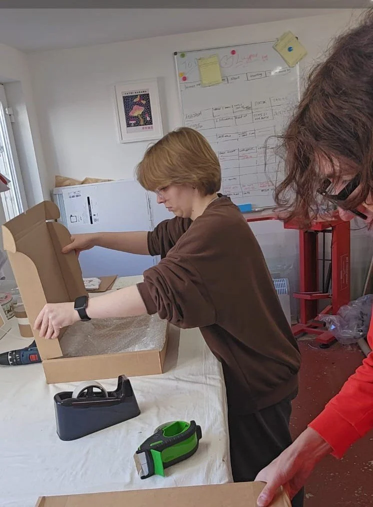 Two people are packing items into a cardboard box on a table, with woodworking or crafting tools nearby, in a bright room with a whiteboard and framed art on the wall.