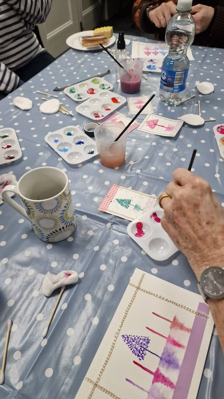 People sit around a table painting watercolor art with palettes, brushes, and paper, with a birthday cake in the background.