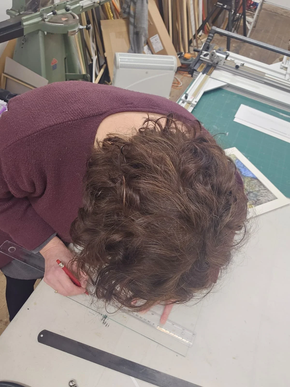 Person with curly brown hair working on a large sheet of paper or fabric, using a ruler and pencil, in a workshop with various tools and frames in the background.