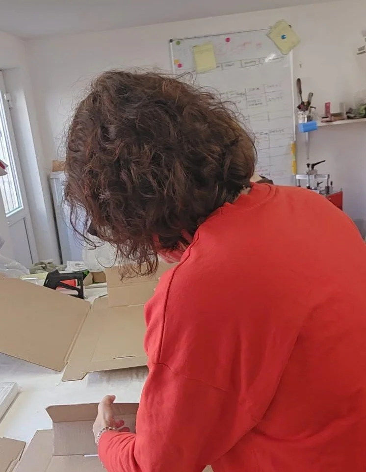 A person with curly hair wearing a red shirt is unboxing cardboard boxes in a room with a whiteboard on the wall.