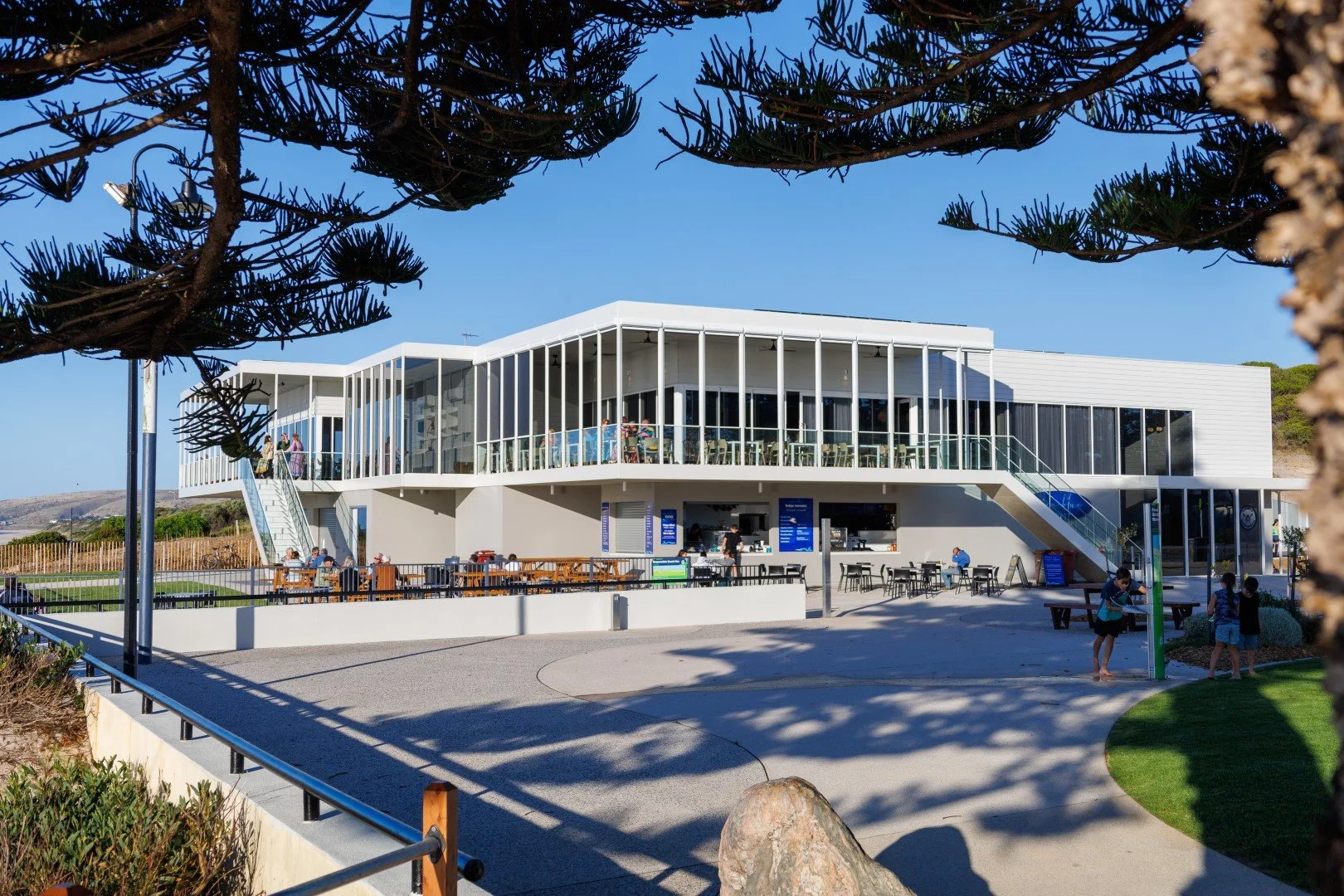 Modern two-story building with large glass windows, outdoor seating area, and people walking and sitting outside, surrounded by trees and clear blue sky.