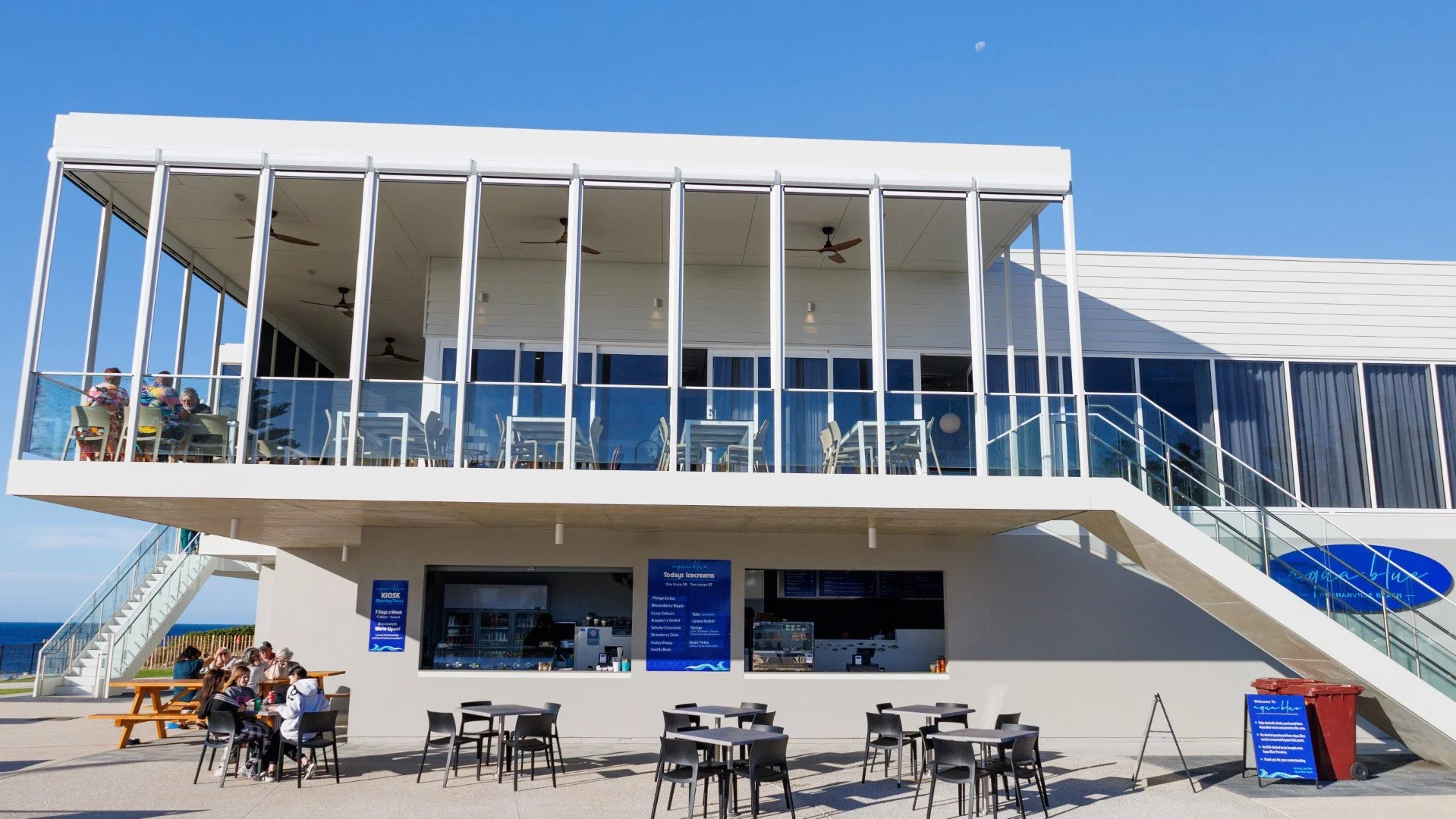 Outdoor view of a modern two-story building with a balcony, glass walls, and an ice cream shop on the ground level. There's an outdoor seating area with tables and chairs, some people sitting, and stairs on both sides leading to the balcony, with a c