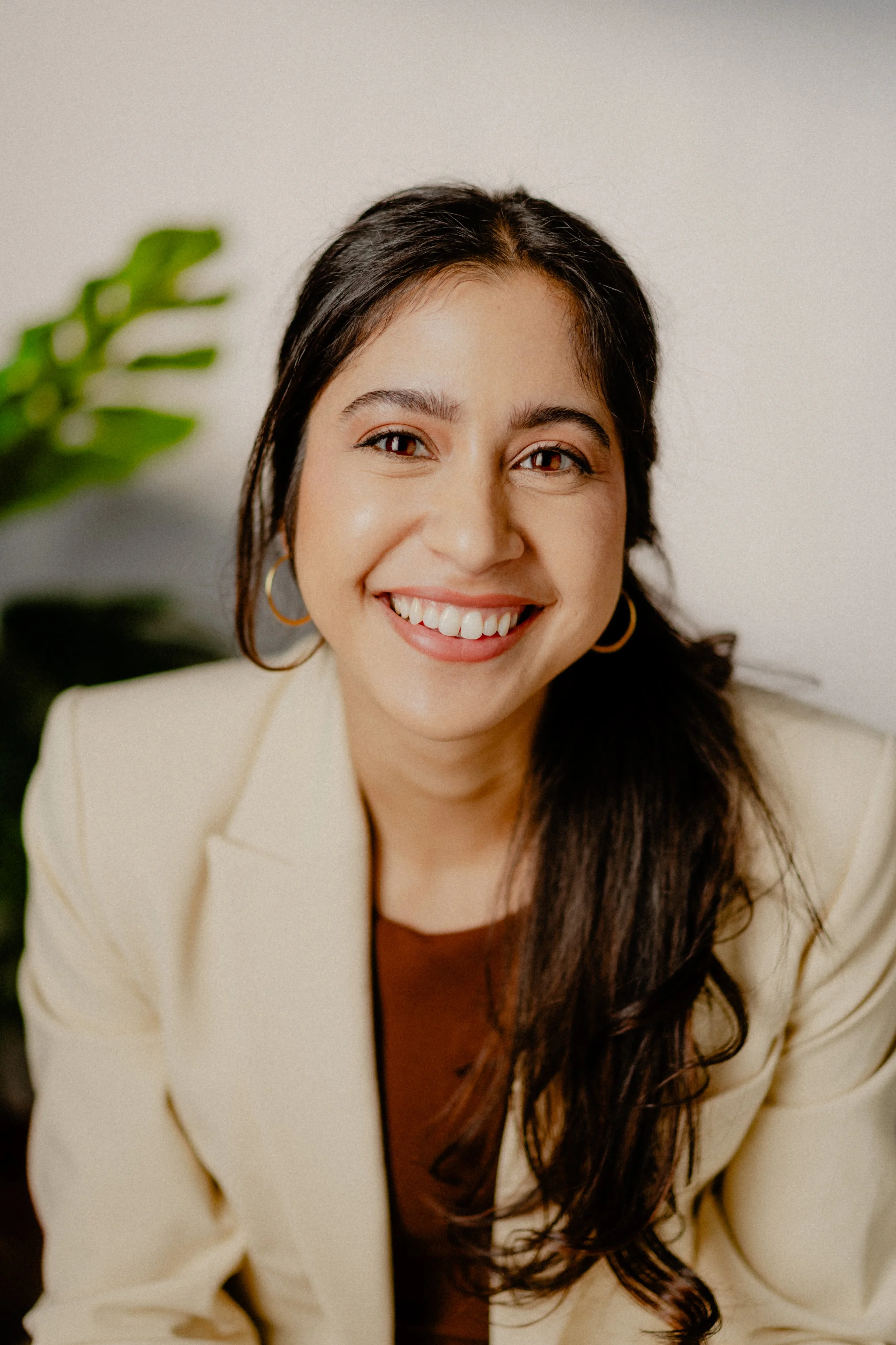 A smiling woman with long dark hair wearing a cream-colored blazer and hoop earrings, sitting indoors with a blurred green plant in the background.