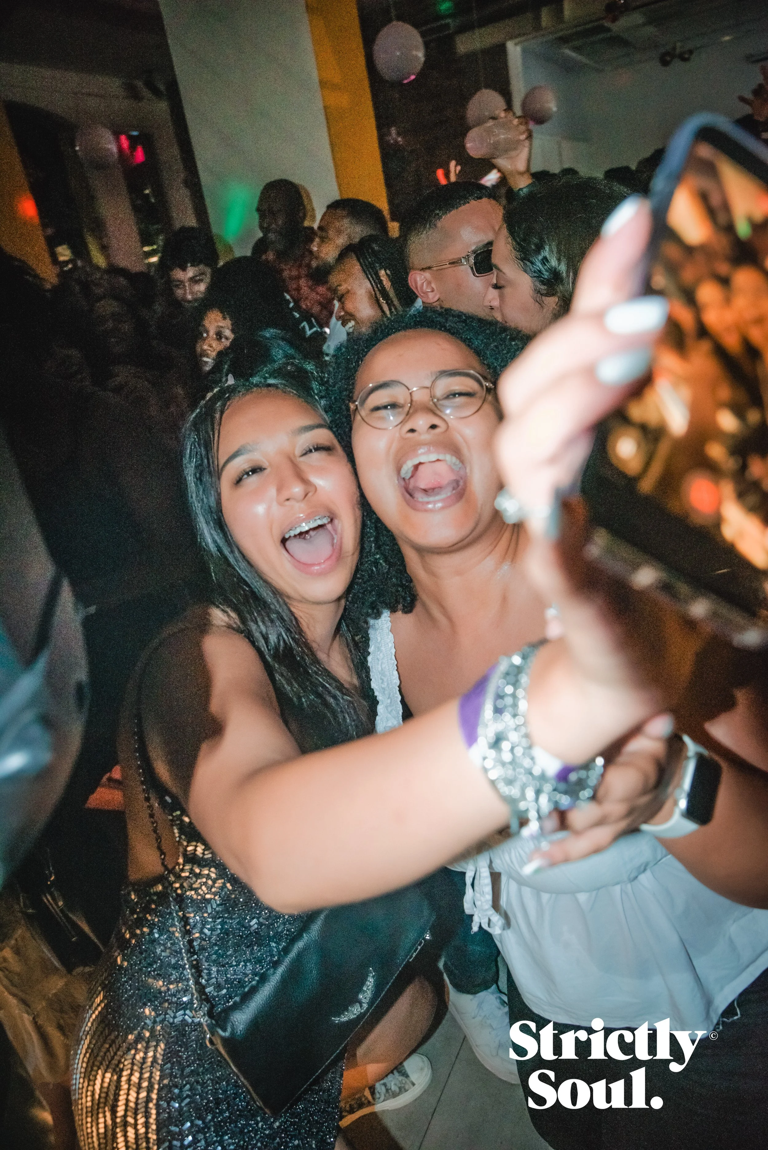 Two women smiling and taking a selfie at a crowded party, with many other people in the background dancing and enjoying themselves.