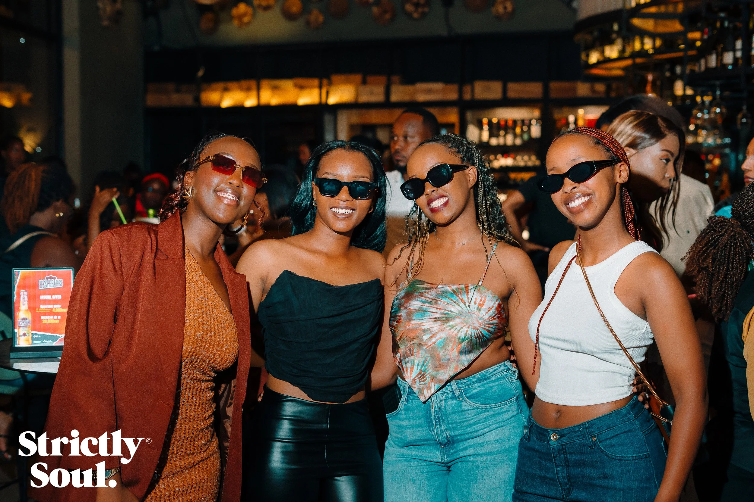 Four women wearing sunglasses and smiling pose together in a lively bar or club environment, with other people and bar shelves in the background.