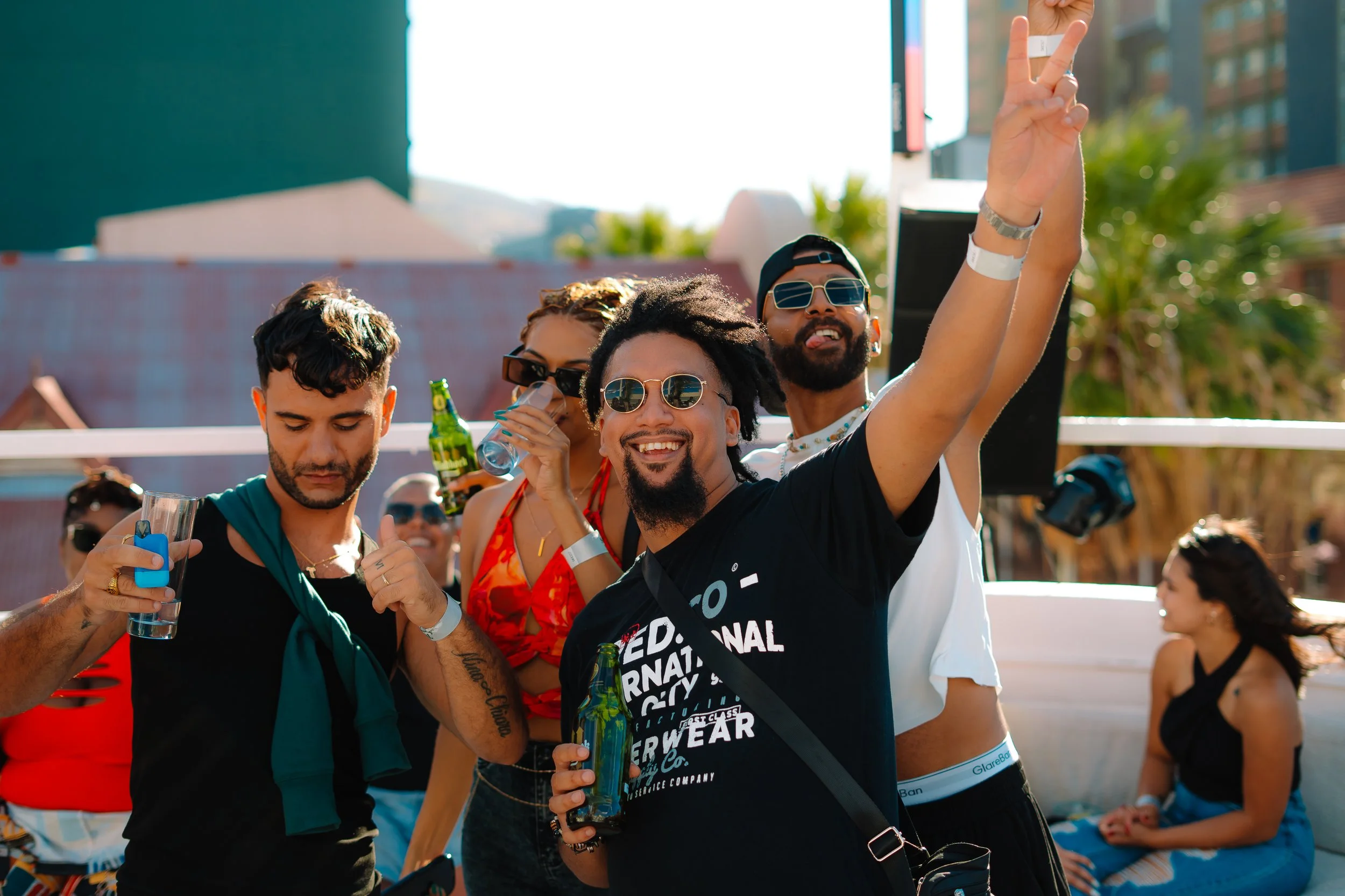 Group of young adults at an outdoor party, smiling and enjoying drinks, with some making peace signs and others relaxed on lounge chairs.
