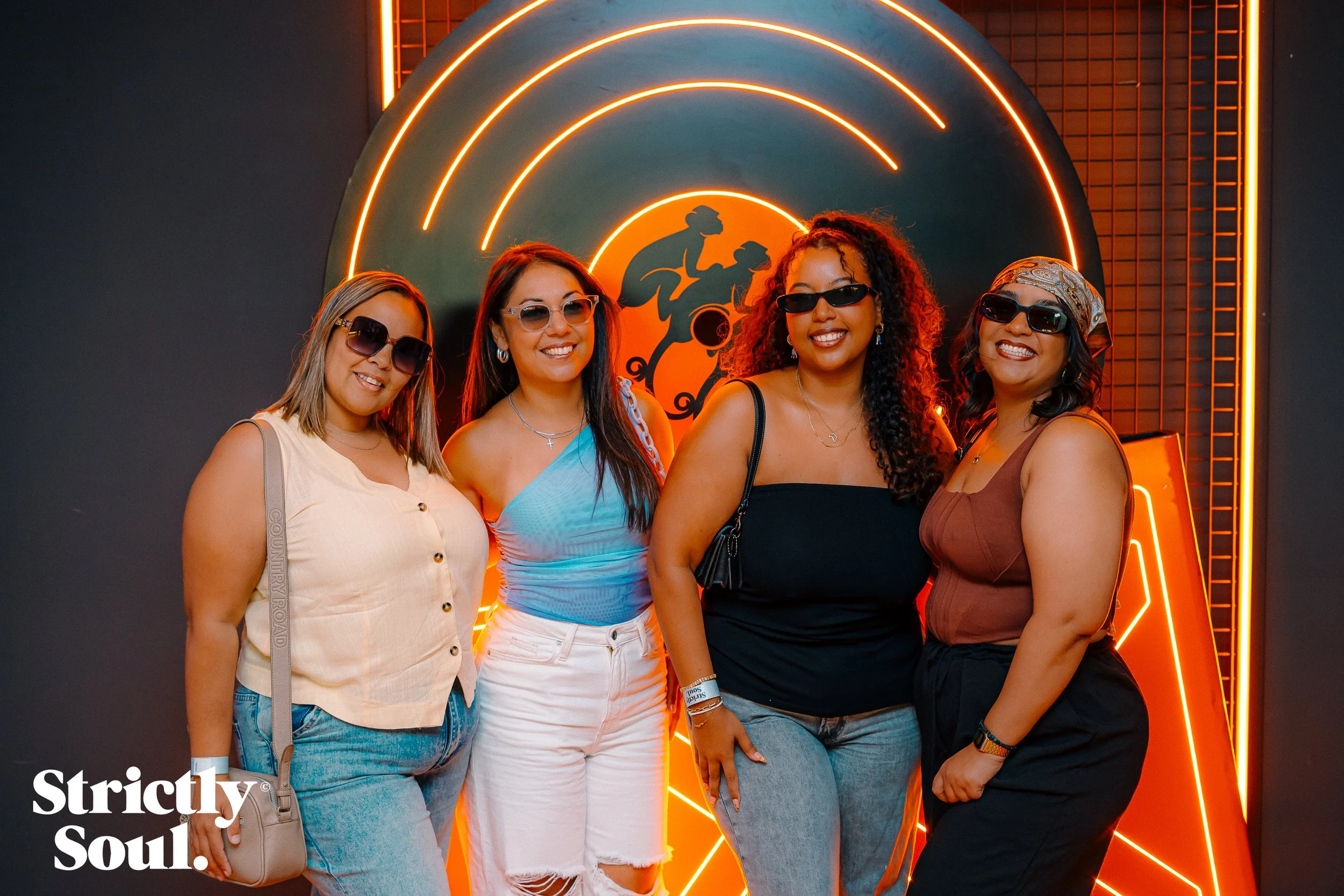Four women wearing sunglasses, smiling, standing in front of a neon-lit artwork with a silhouette of a person riding a motorcycle and the words "Strictly Soul" in the bottom left corner.
