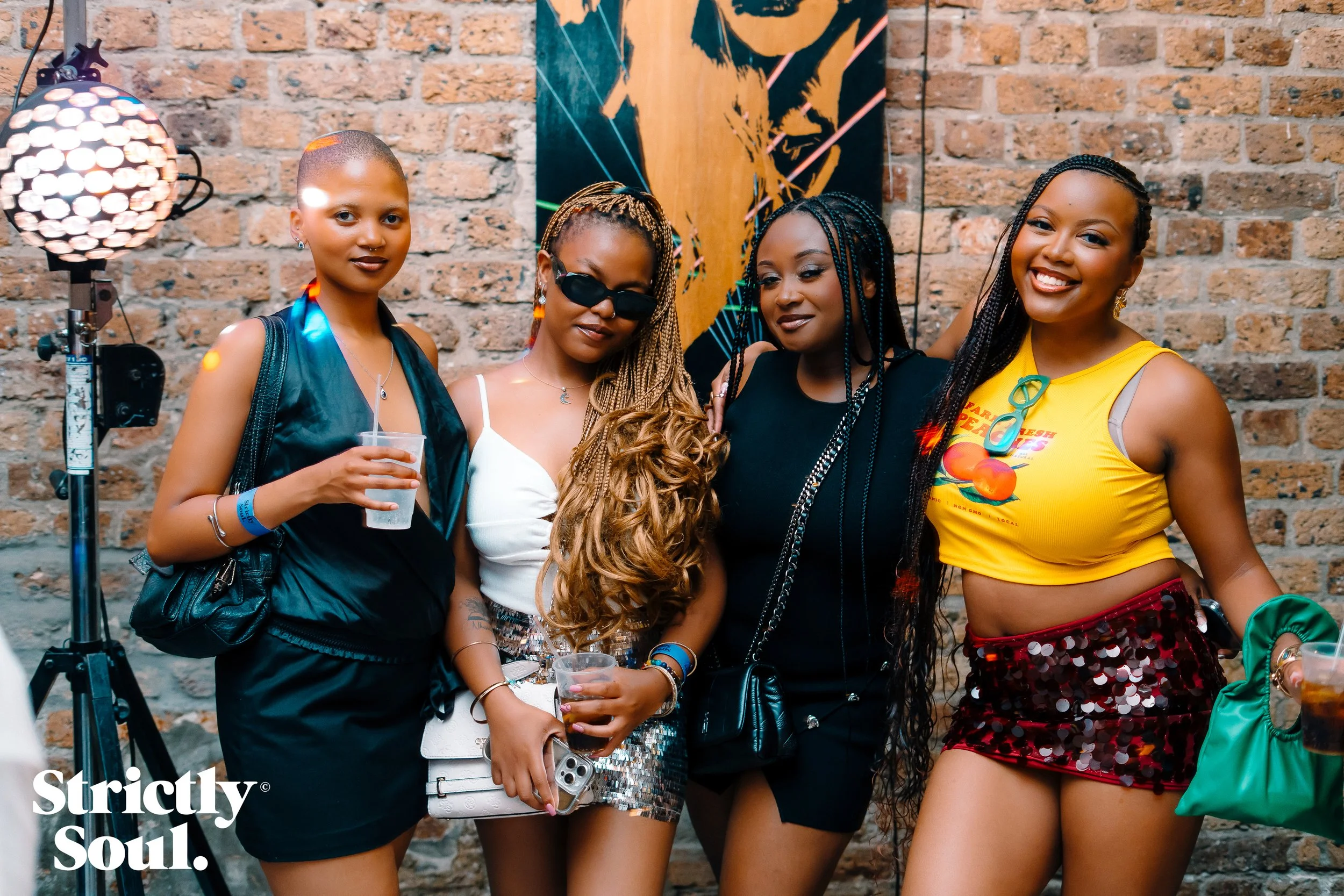 Four women posing together at a social event, standing against a brick wall, with a piece of abstract art and a lamp in the background. They are dressed in stylish, casual attire, holding drinks, and smiling.