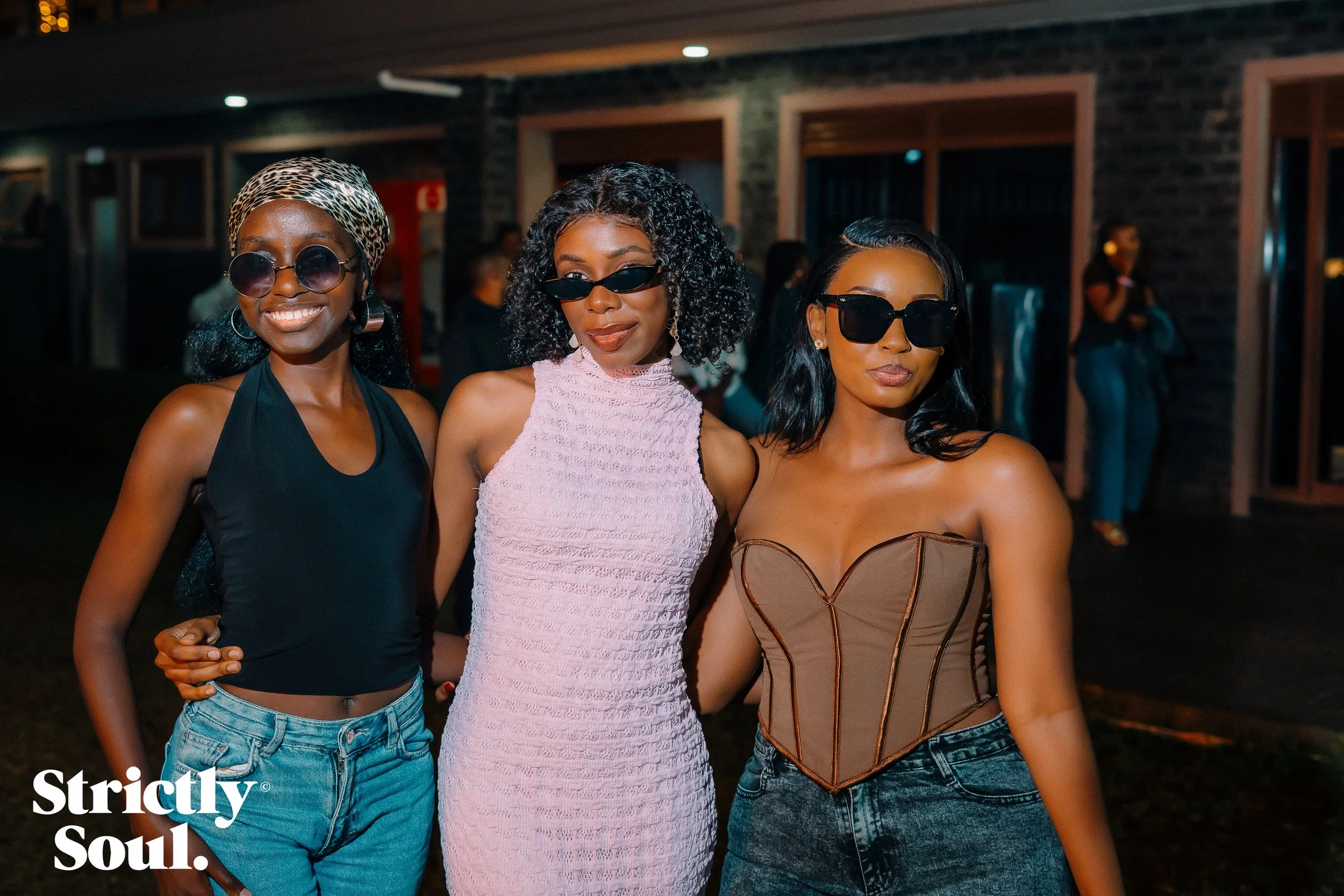 Three women standing close together at night, all wearing sunglasses and stylish outfits, with a brick building and other people in the background.