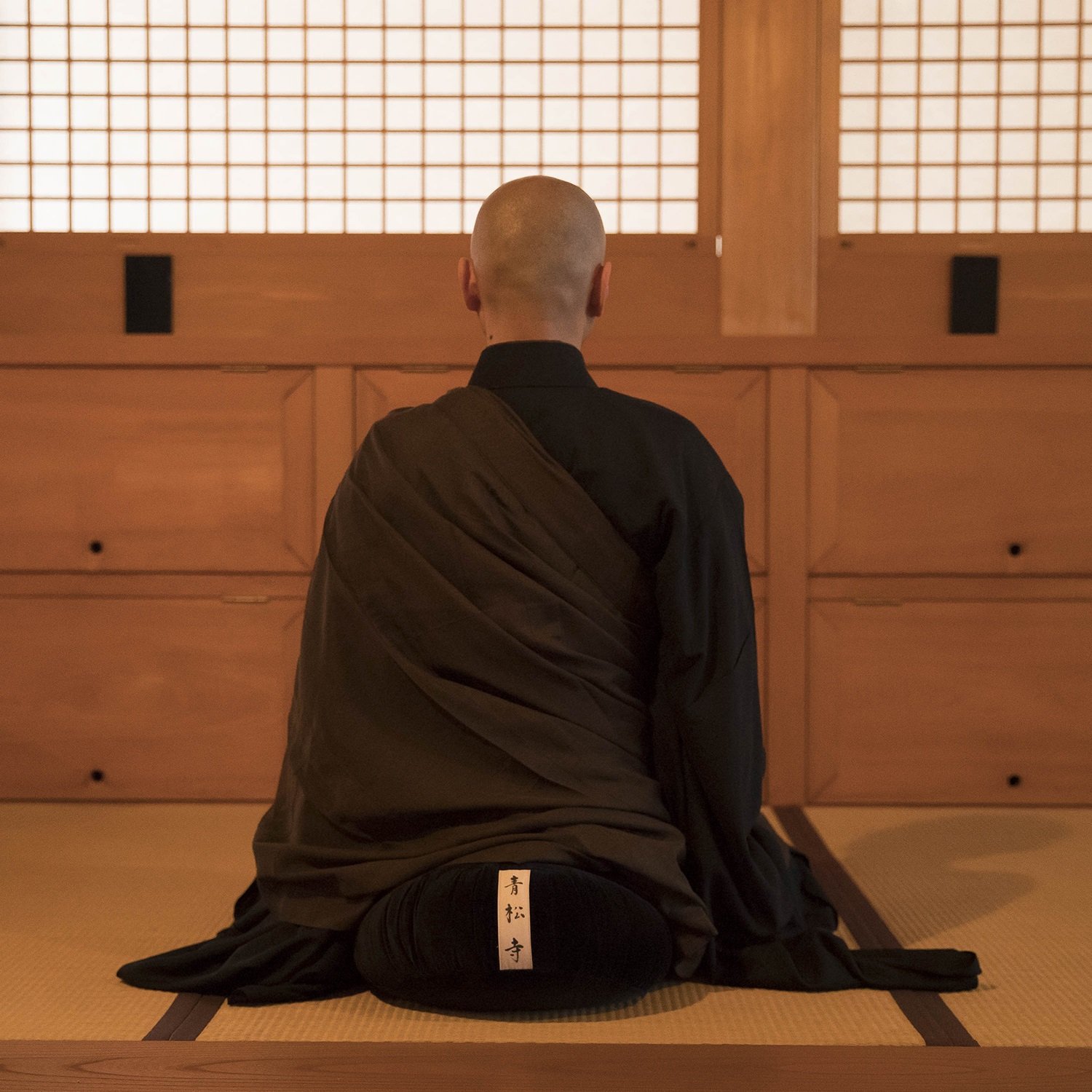 Monje budista en posición de meditación en un templo japonés, con vestimenta tradicional negra y plegaria escrita en un pergamino en su ropa, sentado sobre tatami de madera.