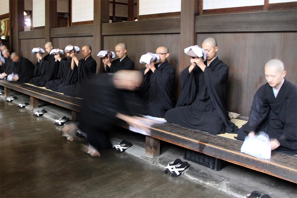 Personas con vestimenta negra sentadas en una estructura de madera, muchas de ellas con la cabeza inclinada hacia sus manos cubriéndose la cara, mientras un monje en movimiento pasa frente a ellas en el interior de un templo tradicional japonés.